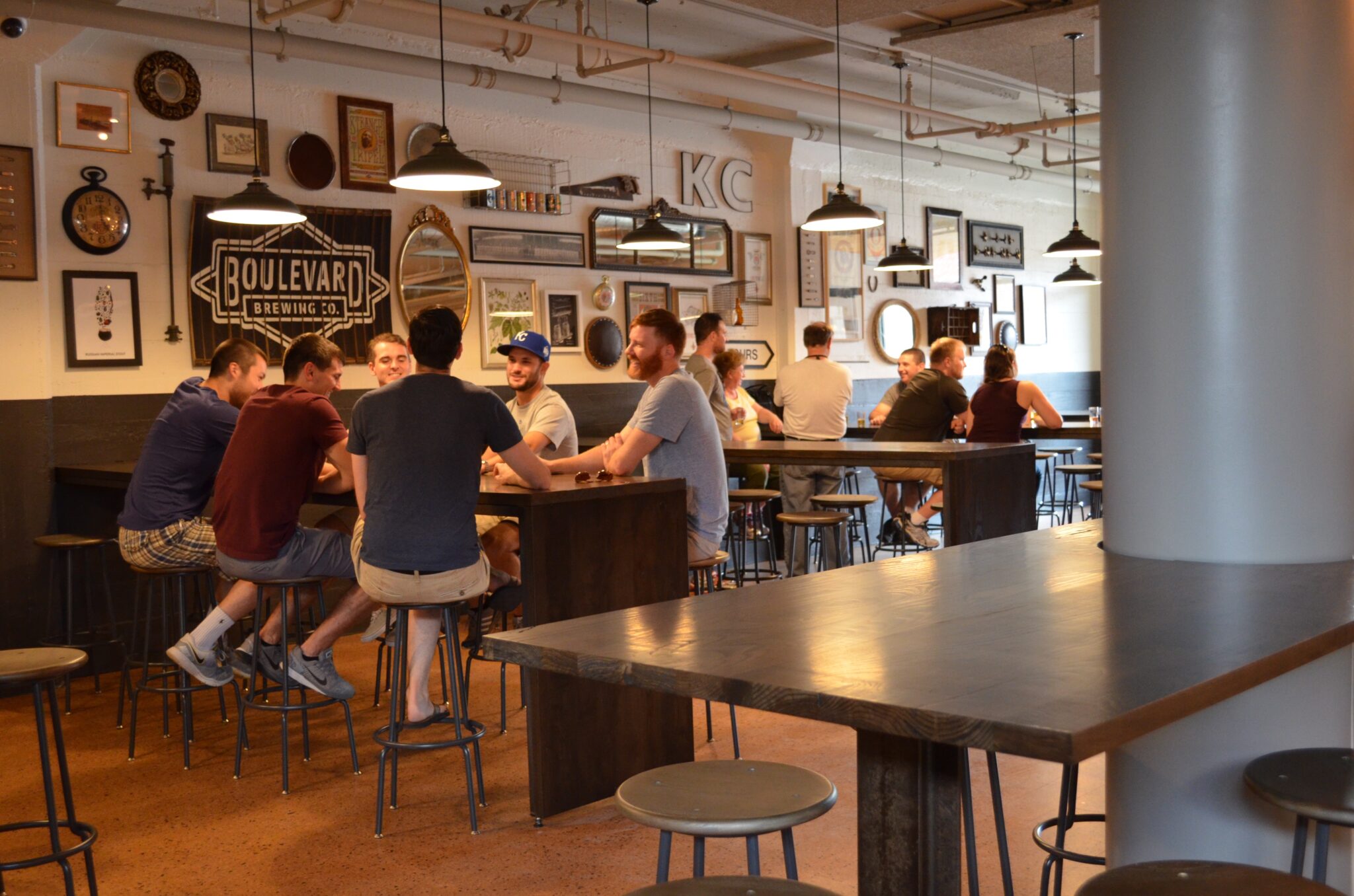 A cozy lounge area with dark leather sofas and small round tables with chairs against a gray wall. A large TV is mounted on the wall above a shelf holding board games. A wooden barrel serves as a table. Shelves display various beer bottles.