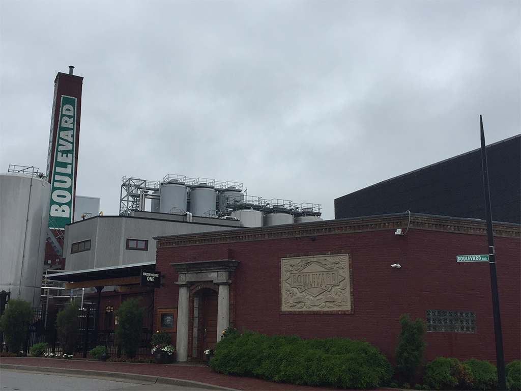 Industrial building with red-brick facade featuring a sign reading "Boulevard." Adjacent is a tall structure with a vertical "Boulevard" sign. Metal tanks are visible above the roof, under a cloudy sky. Street sign at the corner says "Boulevard.
