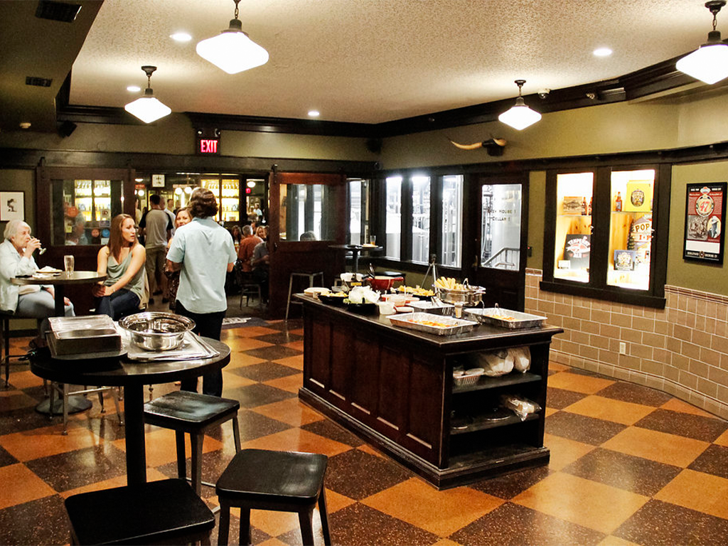 People socialize in a dimly lit restaurant with a buffet table in the center. Some diners sit at tables, while others stand near the buffet. The room has a tiled floor, cushioned chairs, and decorative lighting.