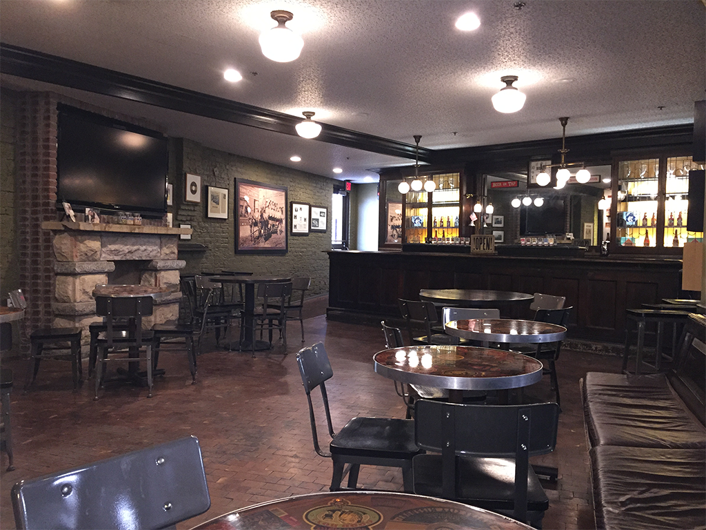 A cozy pub interior with dim lighting, wooden bar, and round tables surrounded by chairs. Walls are decorated with framed pictures, and there's a mounted TV above a stone fireplace. The floor is brick, and the atmosphere is rustic and inviting.