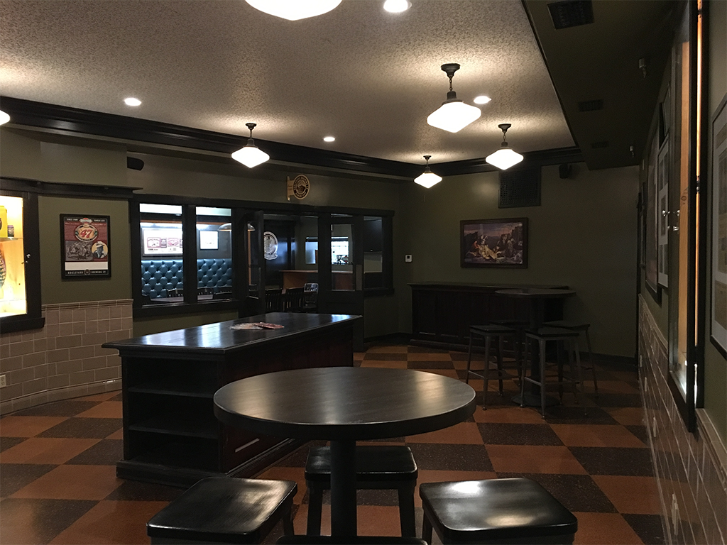 A pub interior with round tables and stools. The lighting is dim with overhead lights. Walls are adorned with framed pictures and posters. The floor has a checkered pattern. A booth with upholstered seating is visible in the background.