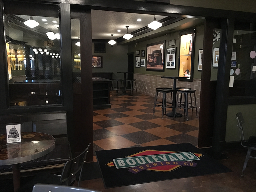 Interior of a bar with checkered flooring, framed photos on green walls, and a "Boulevard Brewing Co" mat on the floor. Several black chairs and tables are placed throughout the dimly lit space.