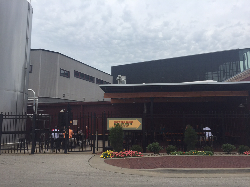 A brewery building with a black fence in front and a sign that reads "Brewery Tours." There are a few people near the entrance, a patio area with tables and umbrellas, and flower beds along the sidewalk.