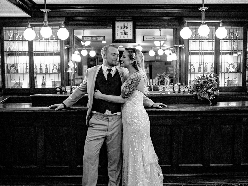 A bride in a lace gown and a groom in a suit and vest stand together, embracing in front of a wooden bar adorned with hanging lights. They are smiling at each other, and a floral arrangement is on the bar. The scene has a vintage ambiance.