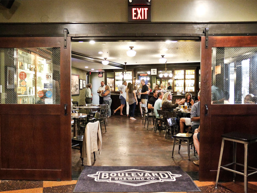 Entrance to a bustling restaurant with a "Boulevard Brewing Co." doormat. People are seated at tables inside, enjoying food and drinks. Large wooden sliding doors are open, and an "EXIT" sign hangs above. Walls feature various decorations and memorabilia.