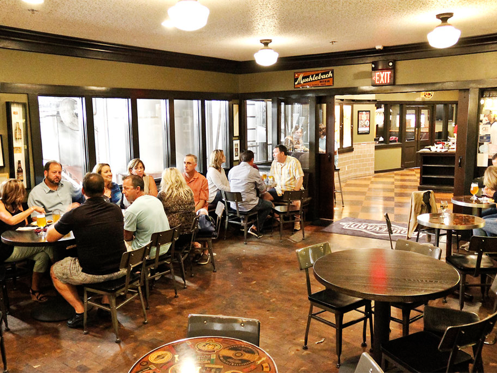 People are gathered in a cozy brewery taproom, sitting at wooden tables and chairs. Some are in conversation, enjoying drinks. The room is warmly lit, with large windows showing brewery equipment in the background. Vintage decor and signs adorn the walls.