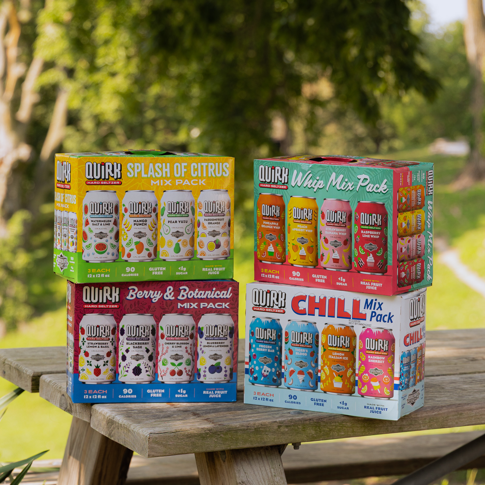 Four colorful Quirk Hard Seltzer mix pack boxes are stacked on a wooden picnic table outdoors, with green trees and a grassy area in the background.