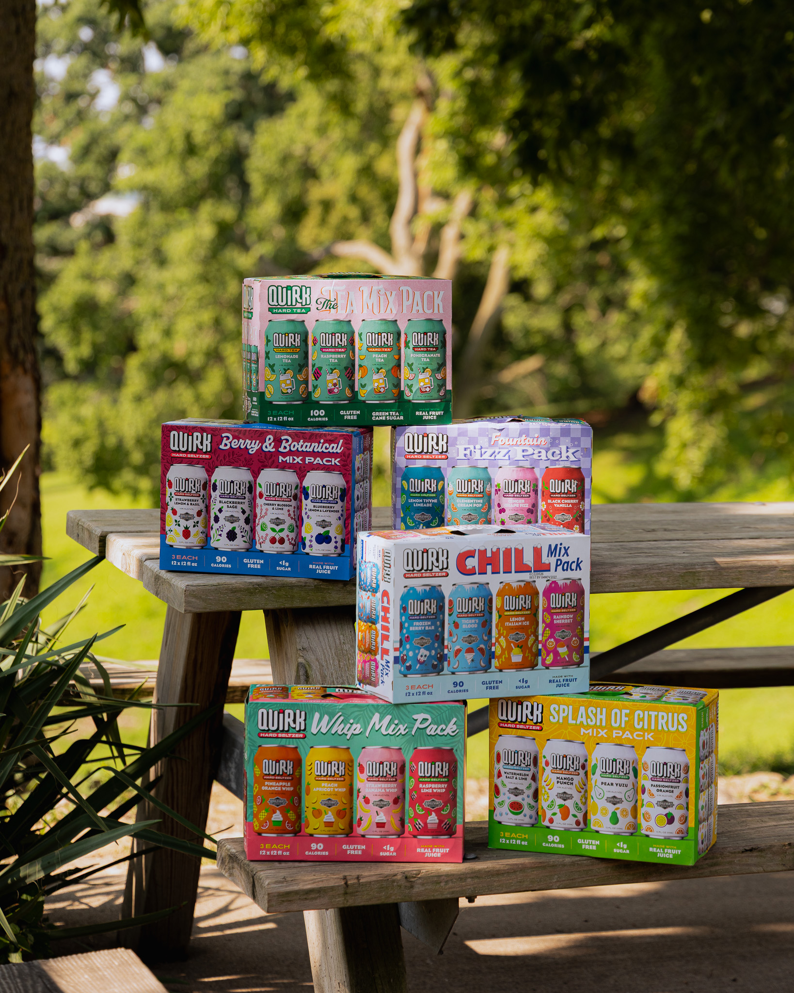 Six colorful boxes of Quirk Hard Seltzer are stacked on a wooden picnic table outside, with green grass and trees in the background. The boxes display various flavors and mix packs.