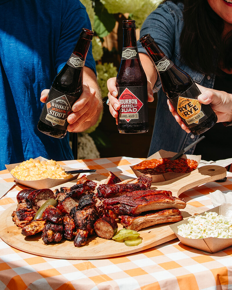 Three people holding dark beer bottles above a wooden platter filled with barbecued meats, pickles, and grilled vegetables. Side dishes like mac and cheese and popcorn sit nearby on an orange and white checkered tablecloth in a sunny outdoor setting.