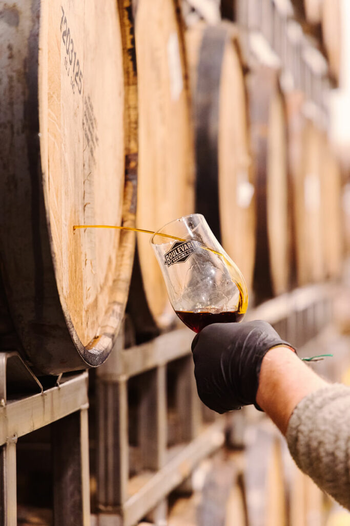 A person in a long-sleeve shirt and black glove is holding a wine glass with red wine under a yellow tube connected to a wooden wine barrel in a storage area. Multiple barrels are visible in the background.