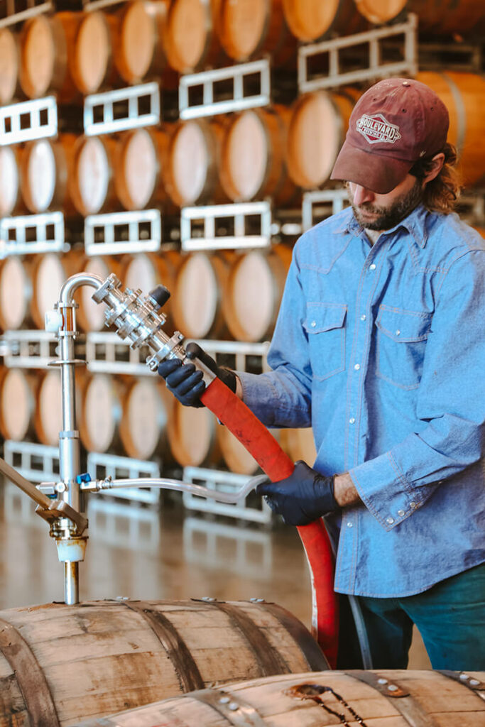 A person wearing a cap, gloves, and a blue shirt is standing in a brewery, using a red hose to fill a wooden barrel. Numerous stacked wooden barrels are visible in the background.