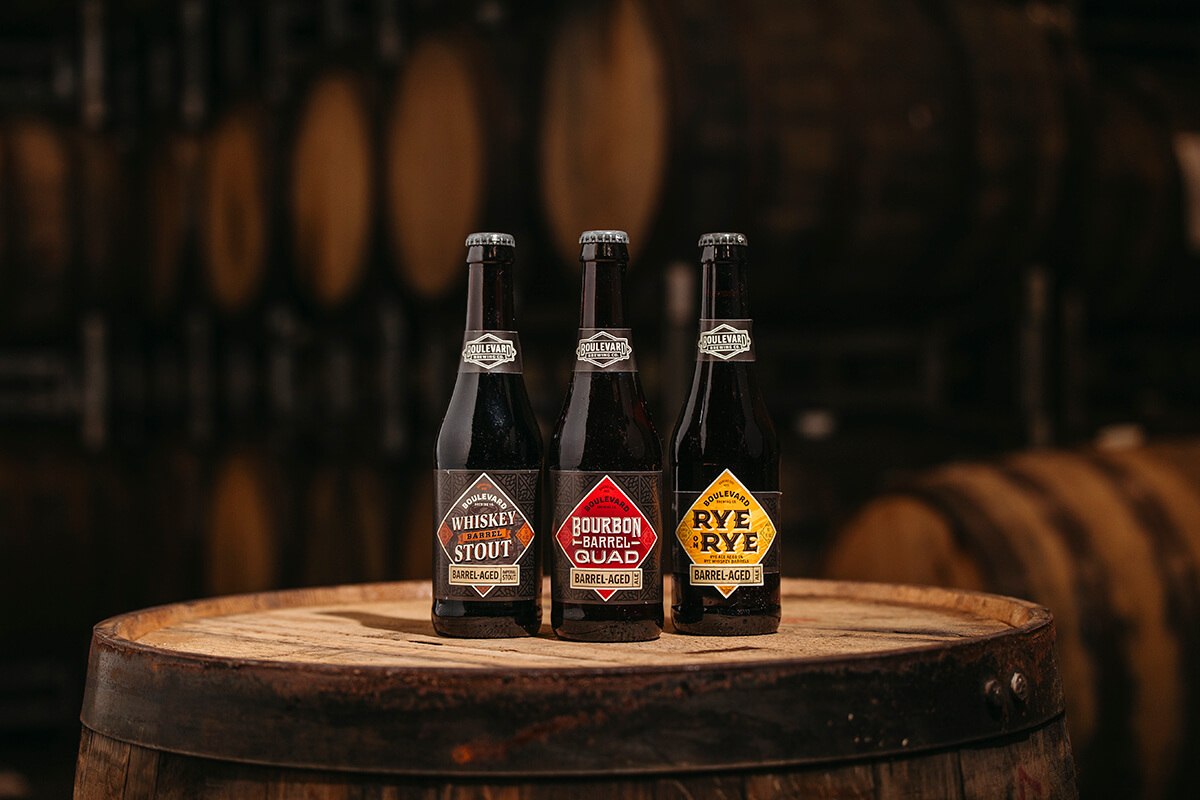 Three bottles of barrel-aged beer, labeled "Whiskey Barrel Stout," "Bourbon Barrel Quad," and "Rye Barrel Rye," are displayed on a wooden barrel. The background features blurred barrels, suggesting a brewery or distillery setting.