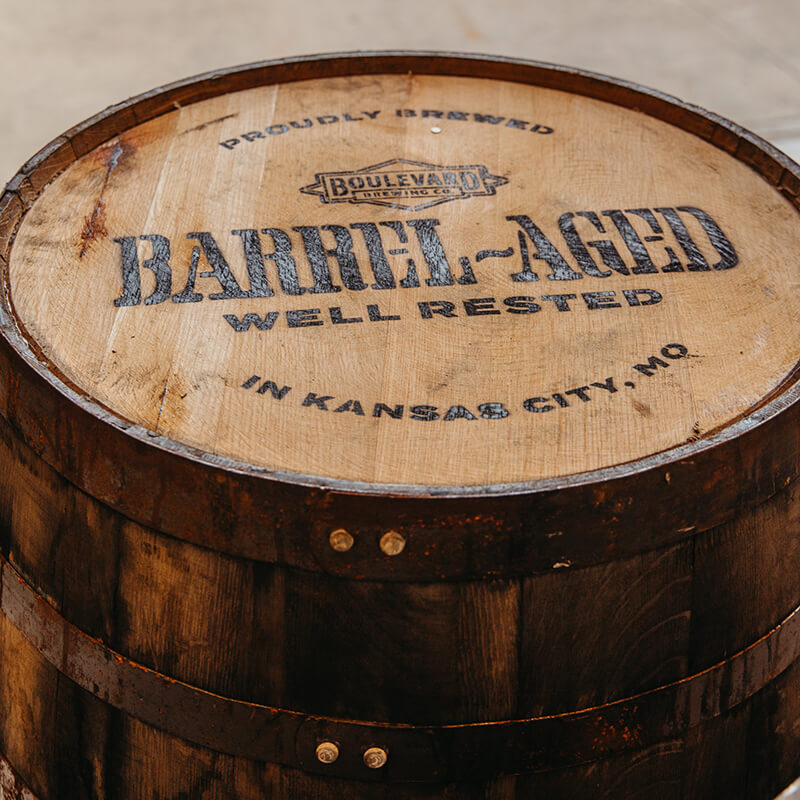 A wooden barrel with the text "PROUDLY BREWED, BOULEVARD BREWING CO., BARREL-AGED, WELL RESTED, IN KANSAS CITY, MO" stamped on its top surface. The barrel is aged and part of a brewing process.