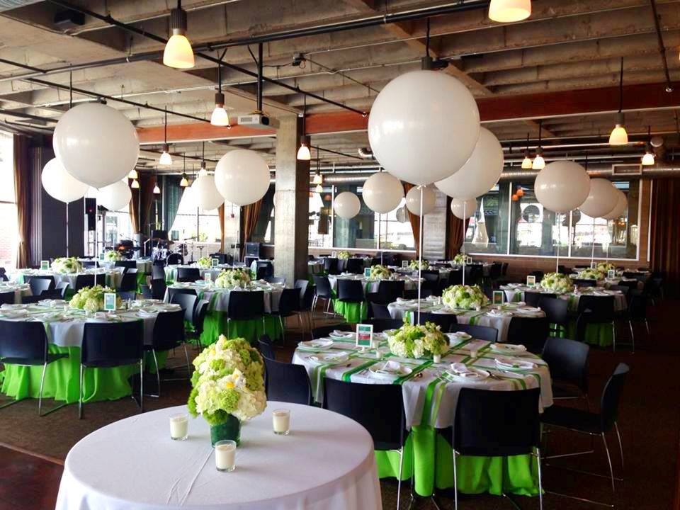 A modern event venue with round tables covered in white and green tablecloths, white floral centerpieces, black chairs, and large white balloons as decorations. The space is well-lit with hanging lights.