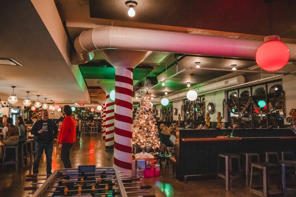 Bar with festive Christmas decor, including a white-lit tree, presents, candy cane-striped column, and round colored lights. People are gathered at tables and standing, creating a lively, holiday atmosphere.