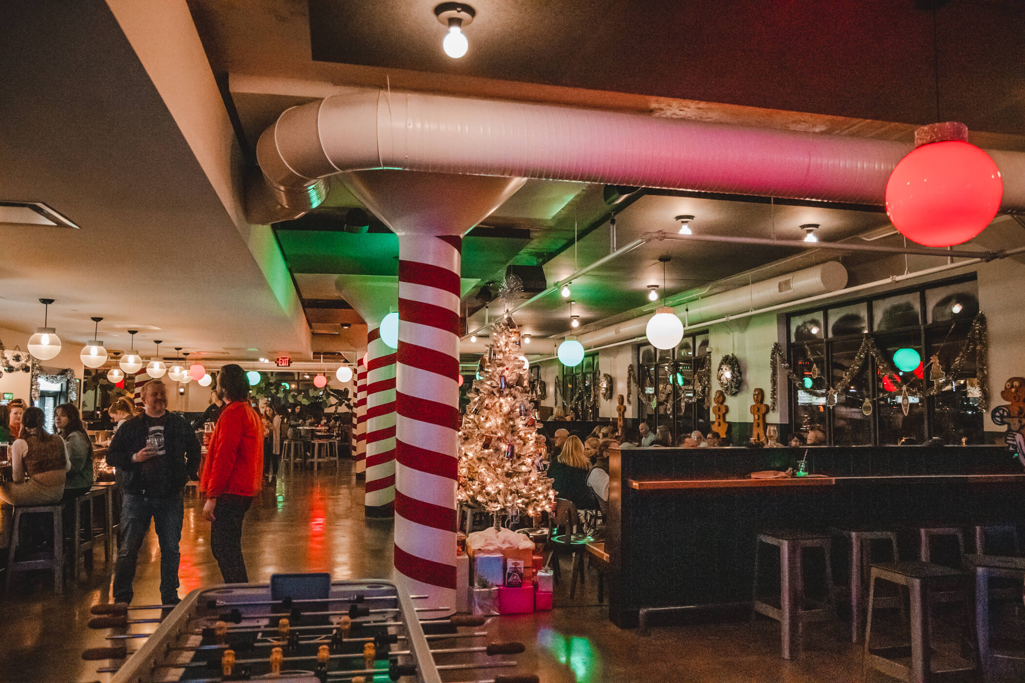 Bar with festive Christmas decor, including a white-lit tree, presents, candy cane-striped column, and round colored lights. People are gathered at tables and standing, creating a lively, holiday atmosphere.