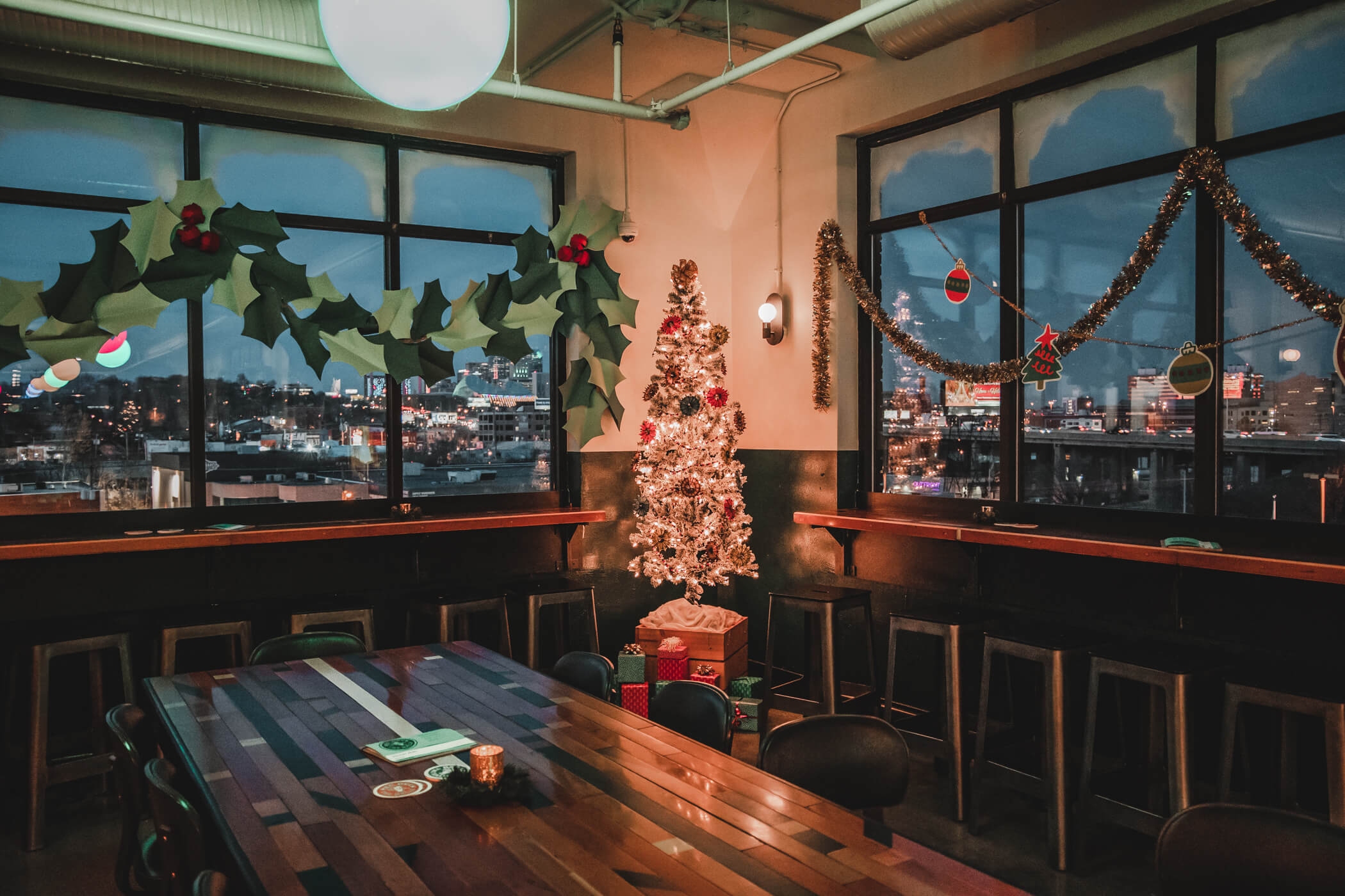 A cozy room decorated for Christmas with garlands, holly, and ornaments. A lit Christmas tree with presents underneath stands by large windows overlooking a cityscape at dusk.