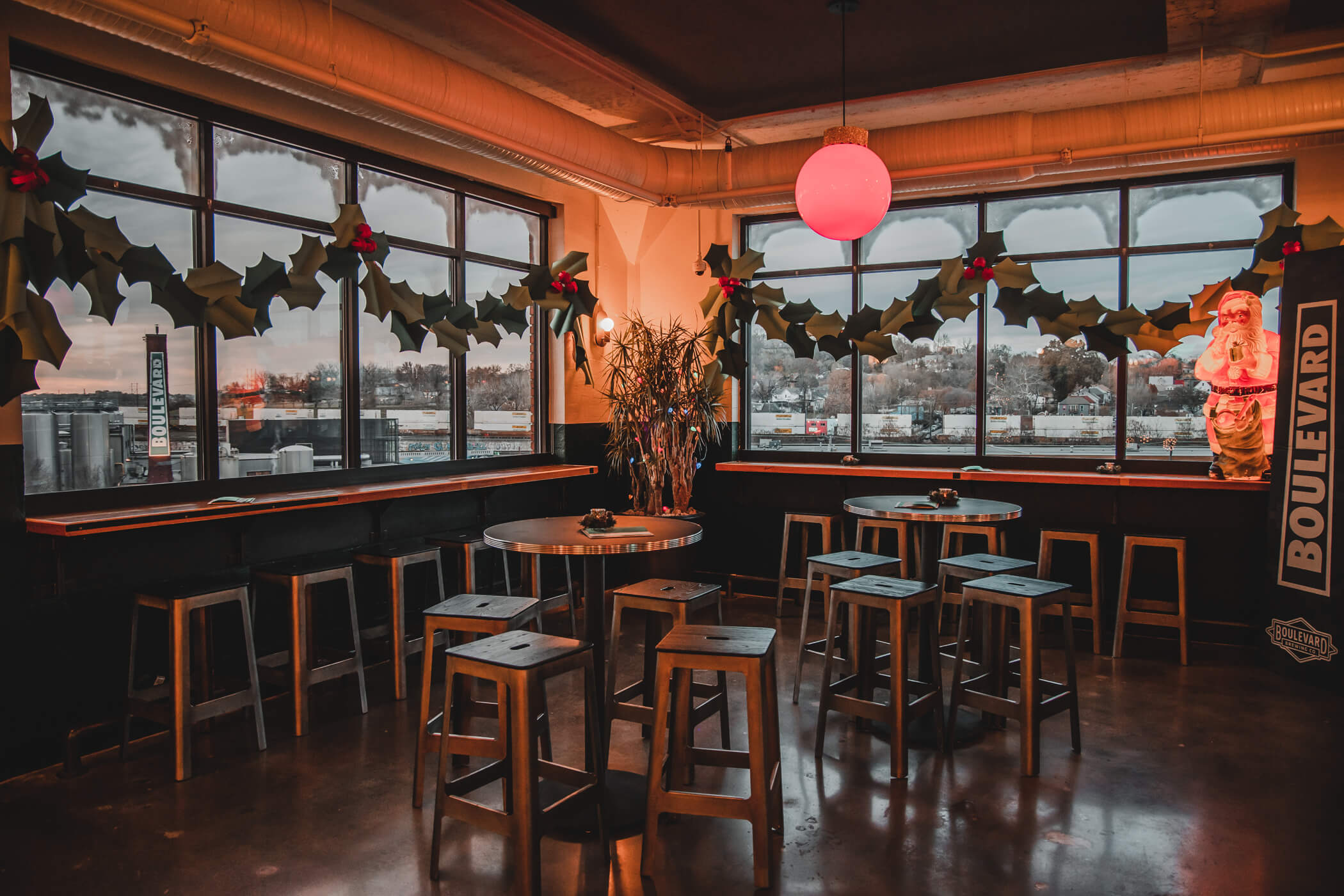 Festively decorated cafe with wooden tables and stools, large windows adorned with holly garlands and a Santa decoration, a glowing red light, and a view of an industrial area outside.