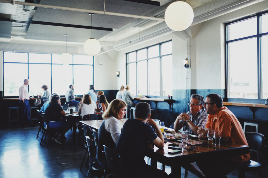 People sit and converse at wooden tables in a bright, modern restaurant with large windows and round hanging lights. Some eat or drink while others chat, creating a casual and lively atmosphere.