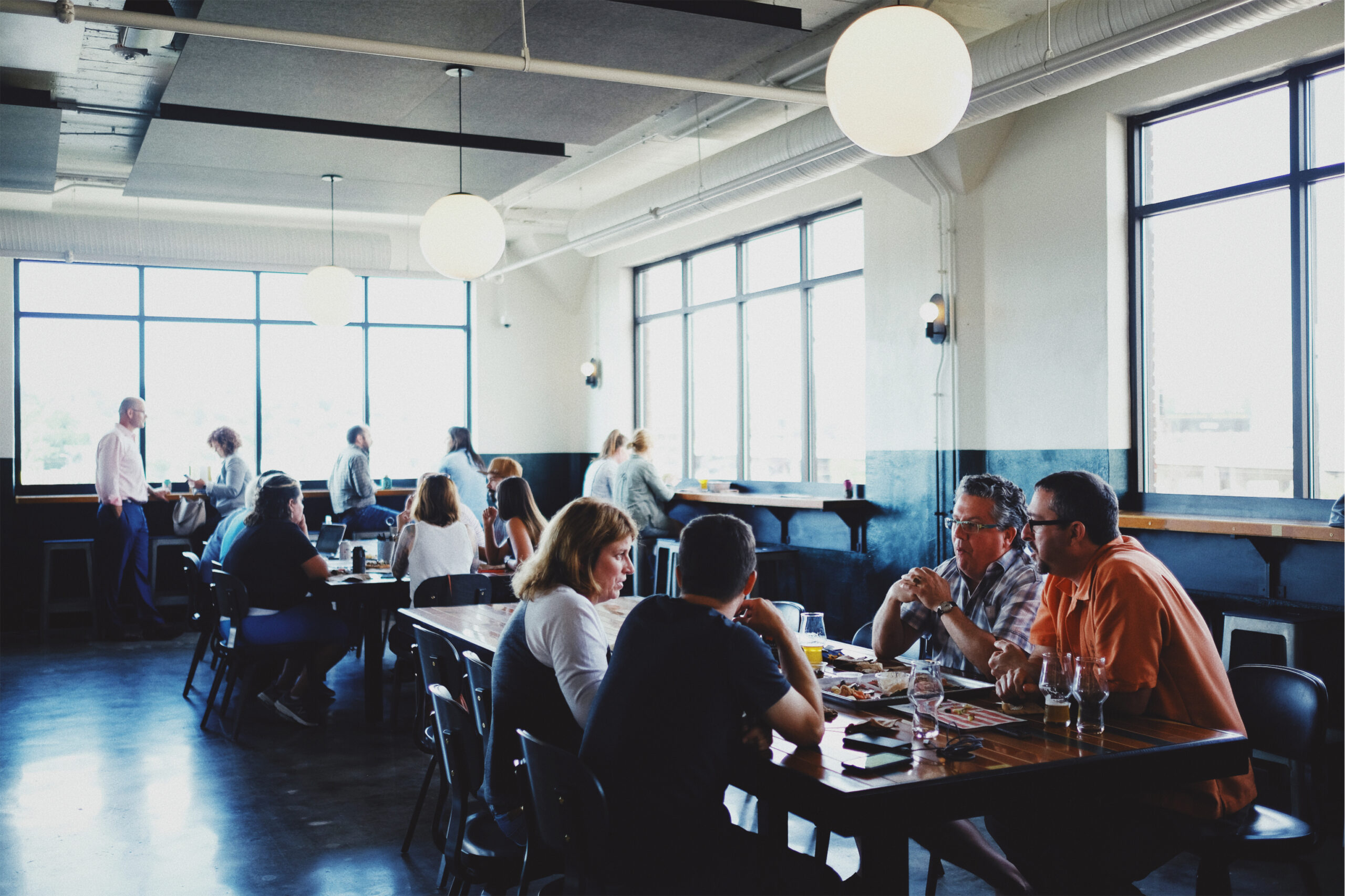 People sit and converse at wooden tables in a bright, modern restaurant with large windows and round hanging lights. Some eat or drink while others chat, creating a casual and lively atmosphere.