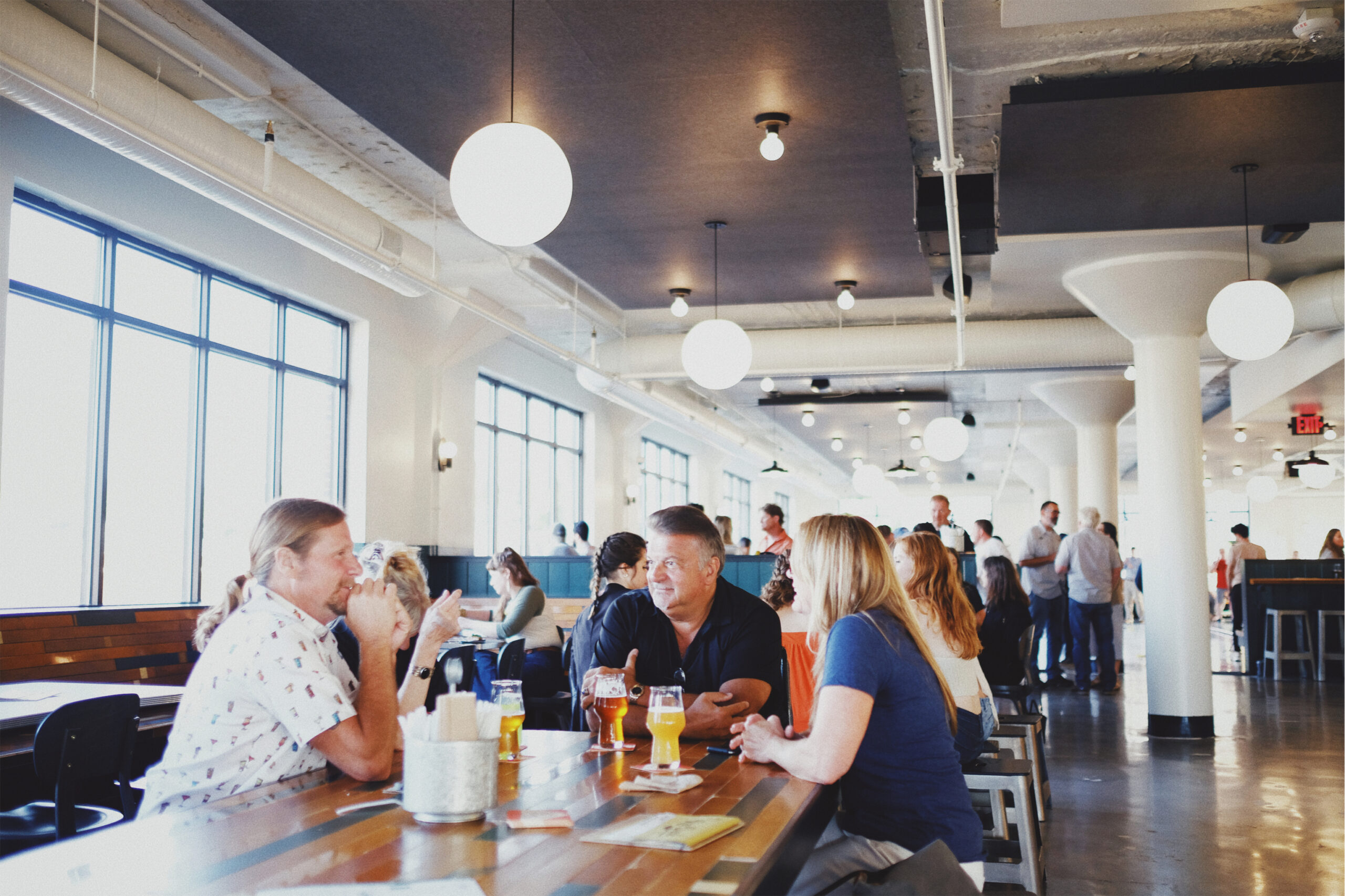 People sit at long wooden tables in a modern, bright, industrial-style restaurant or bar, talking and drinking. Large windows let in natural light, and round ceiling lights hang throughout the space.