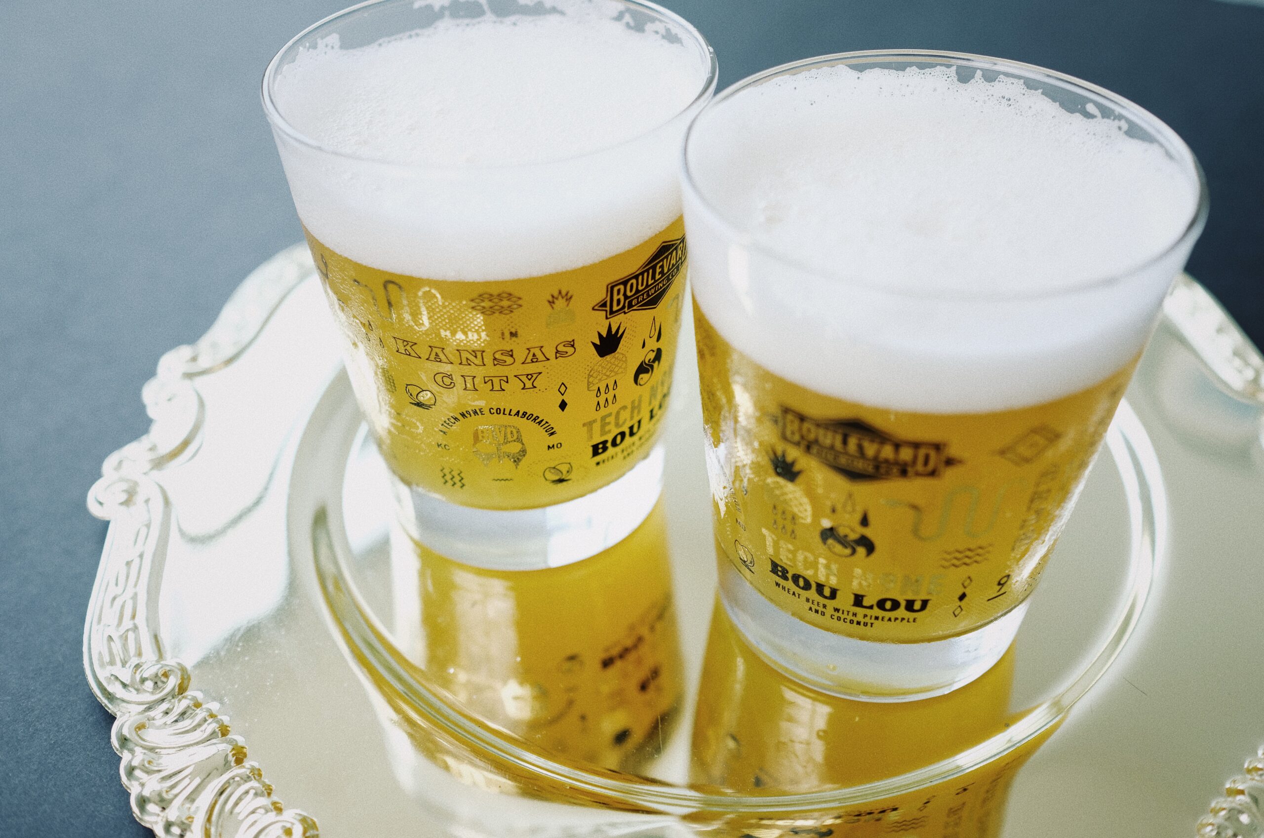 Two frothy glasses of beer on a reflective silver tray. The glasses have decorative designs and text related to Kansas City and Boulevard Brewing Co. The tray features intricate edging, and it's set against a smooth, dark background.