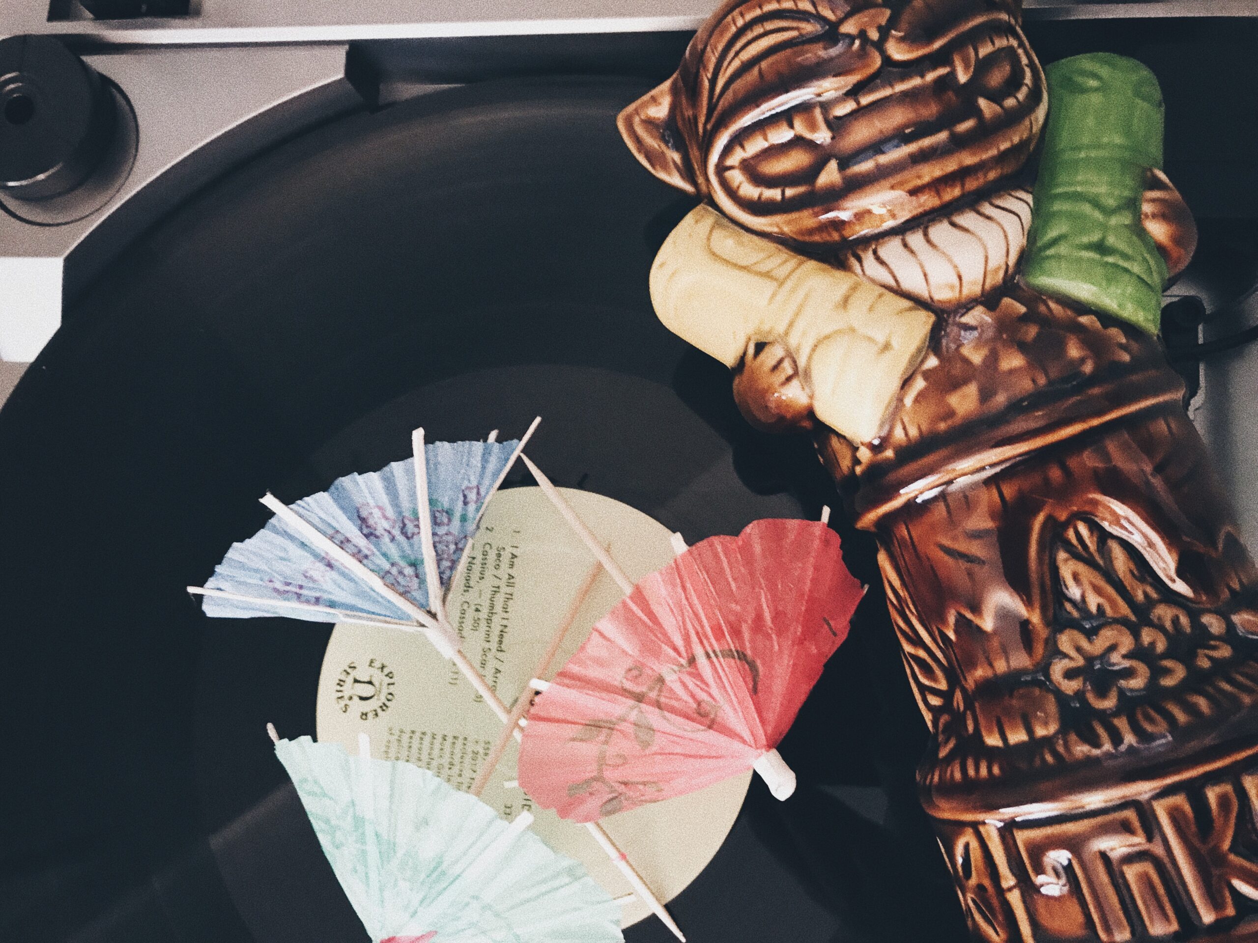 A vinyl record is spinning on a turntable, surrounded by colorful paper umbrellas. There's a decorative tiki mug placed on the record, adding a tropical vibe to the scene.