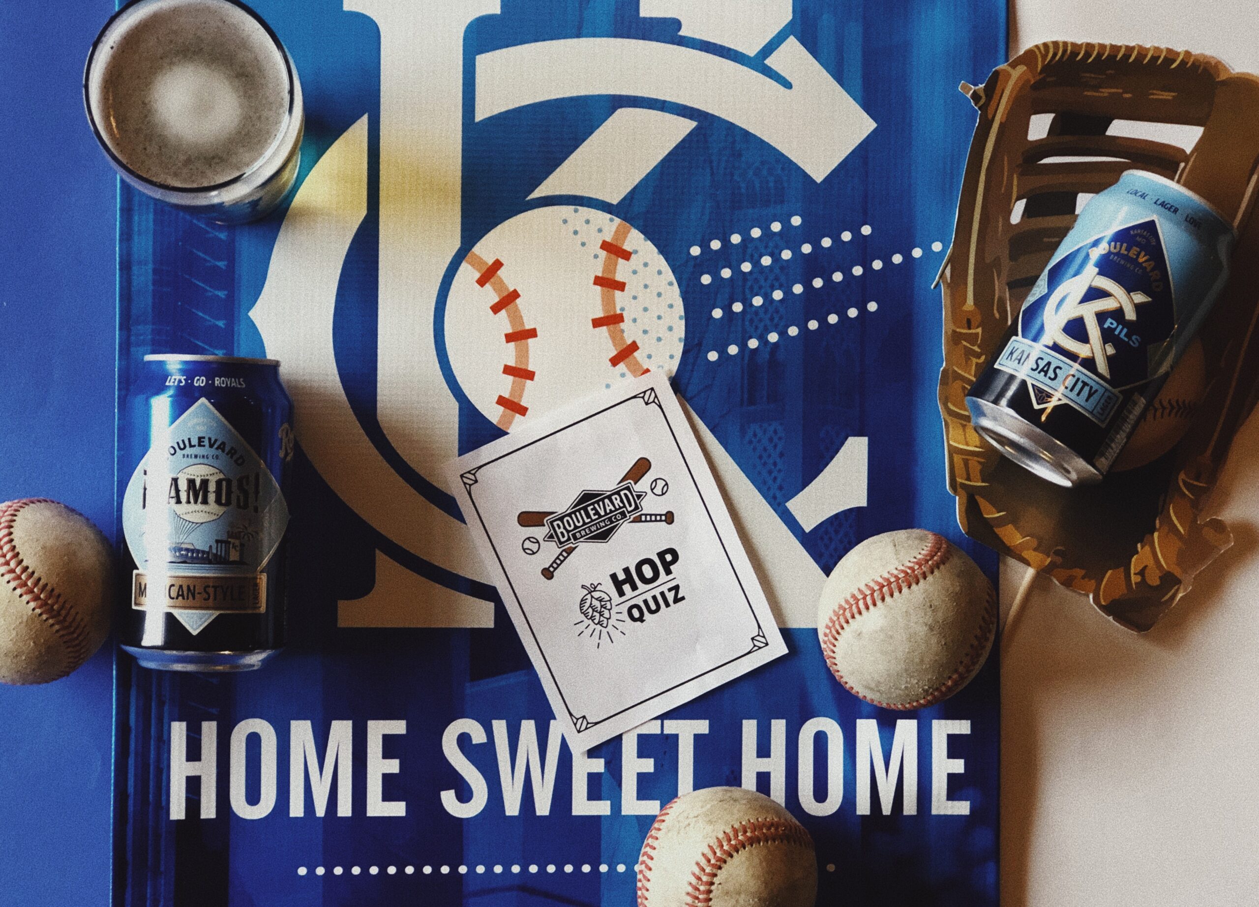 A sports-themed scene featuring baseballs, a glove, two cans of Boulevard beer, and a "Hop Quiz" card on a blue "Home Sweet Home" baseball poster. The design includes baseball stitching and beer details.