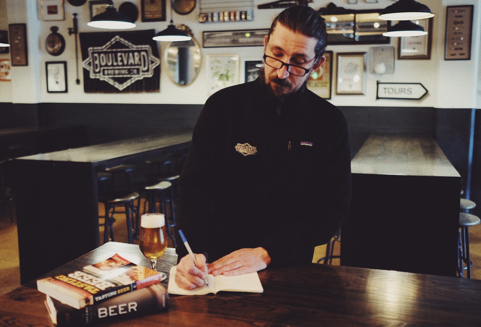 A bearded man with glasses writes in a notebook at a wooden table. A beer glass and two open books, one titled "The Beer Bible," are beside him. The background has framed pictures and industrial-style lights in a cozy bar setting.