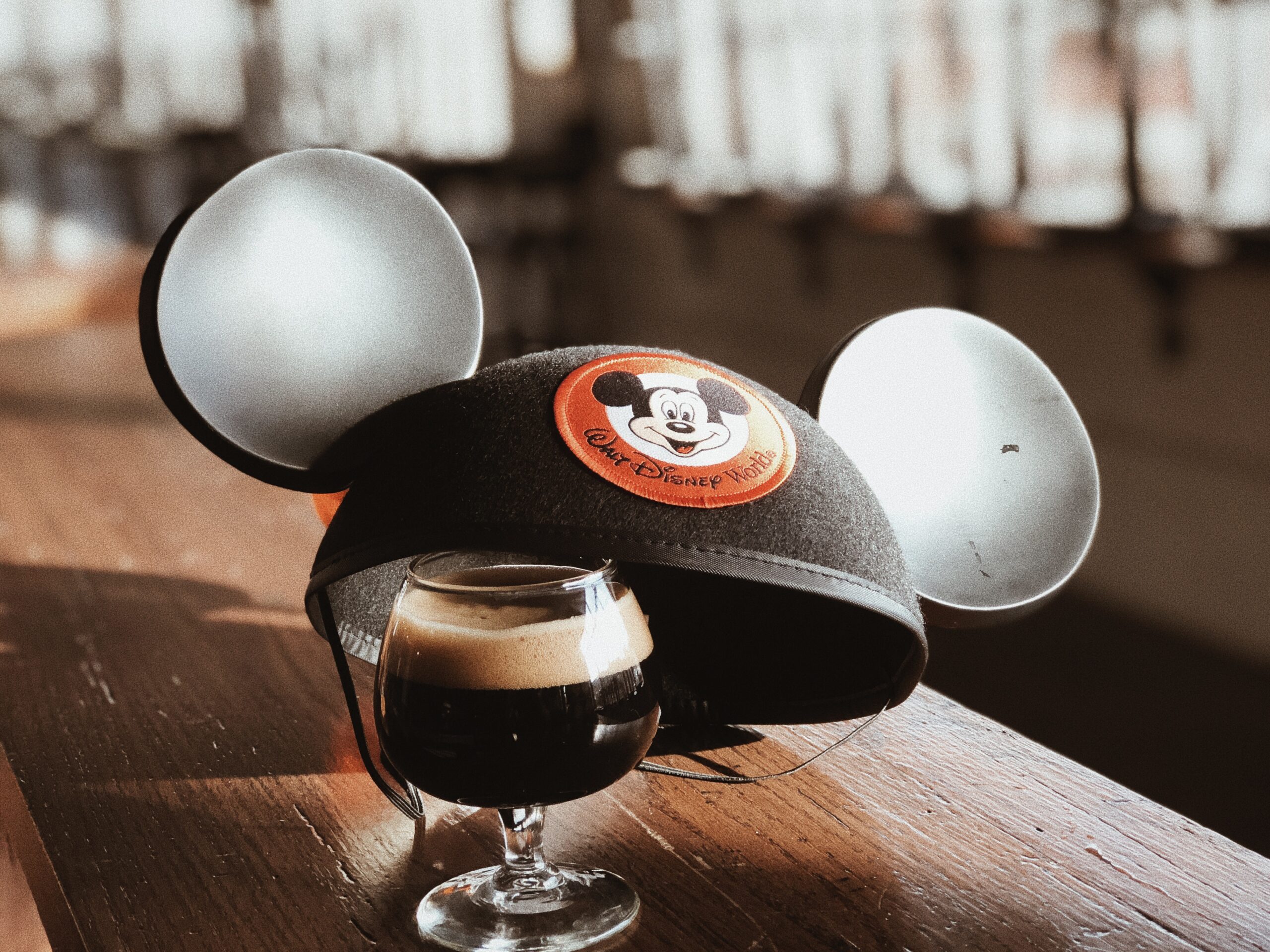 A small glass of dark beer sits on a wooden table with a pair of Mickey Mouse ears beside it. The ears have a "Walt Disney World" emblem featuring Mickey Mouse. Sunlight streams through blurred windows in the background, creating a warm ambiance.