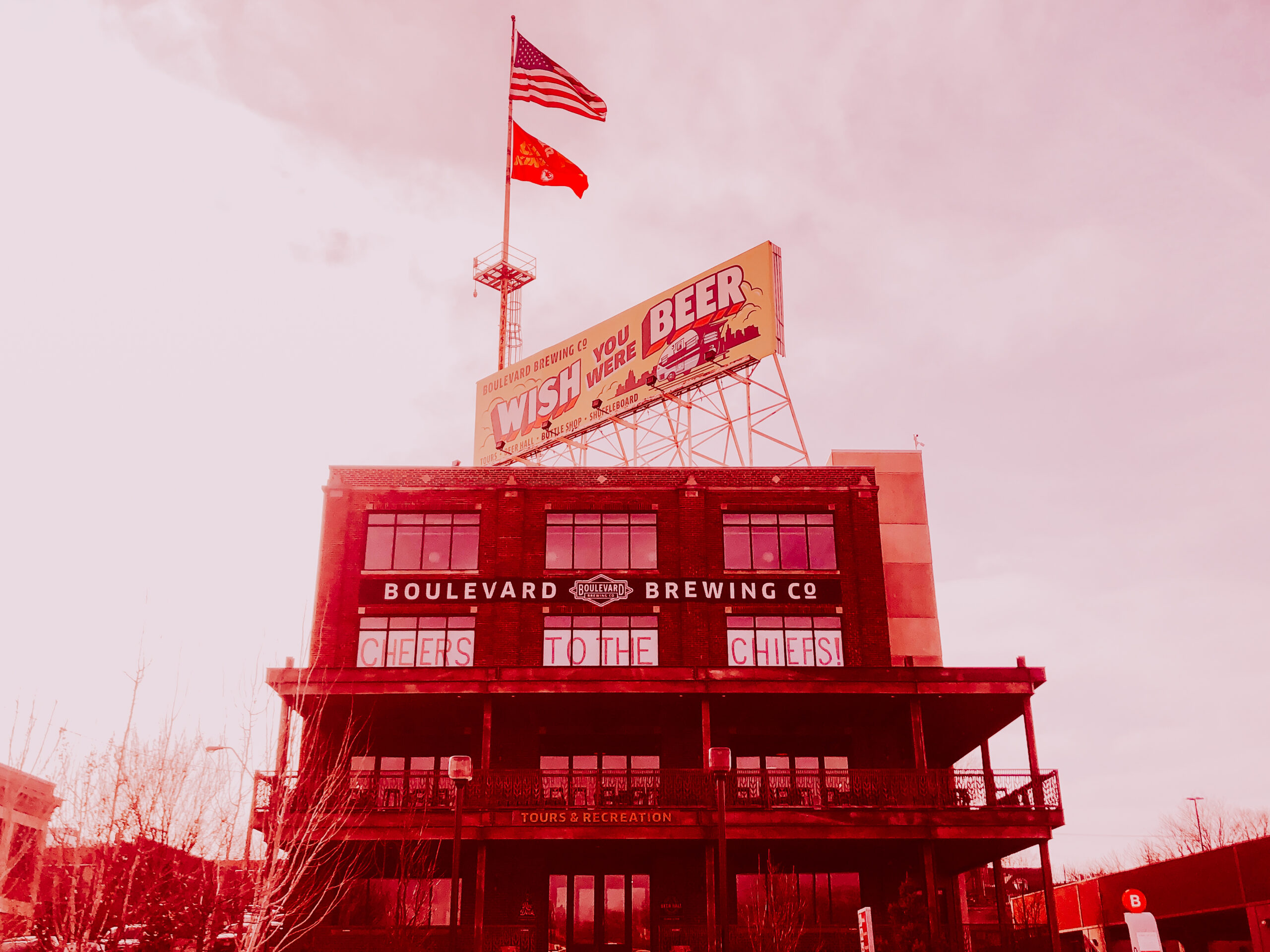 A red-tinted image of a multi-story brick building with an American flag and another flag atop a pole. A large sign reads, "WISH YOU WERE BEER," with "BOULEVARD BREWING CO" below it and "CHEERS TO THE CHEERS" displayed in upper windows.