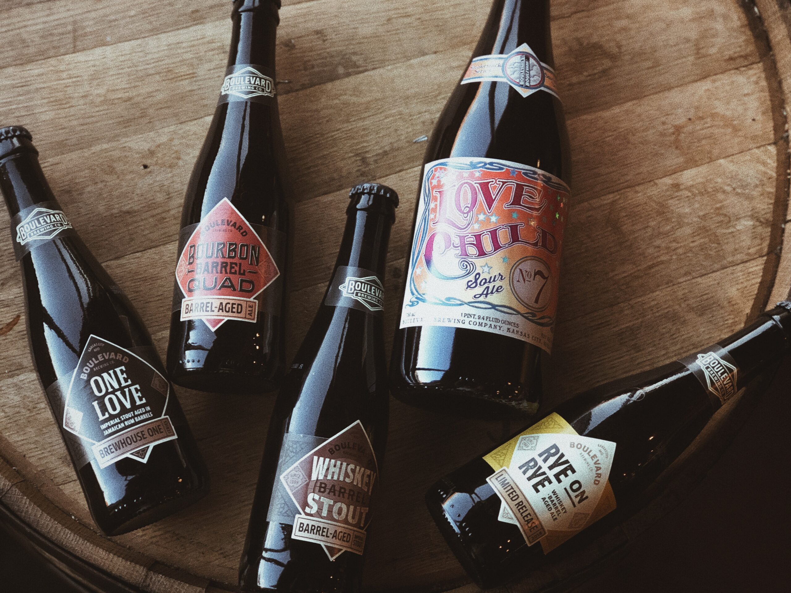 Five assorted craft beer bottles are arranged on a wooden surface. Each bottle has a unique, colorful label and cap, showcasing different types and styles of beer.