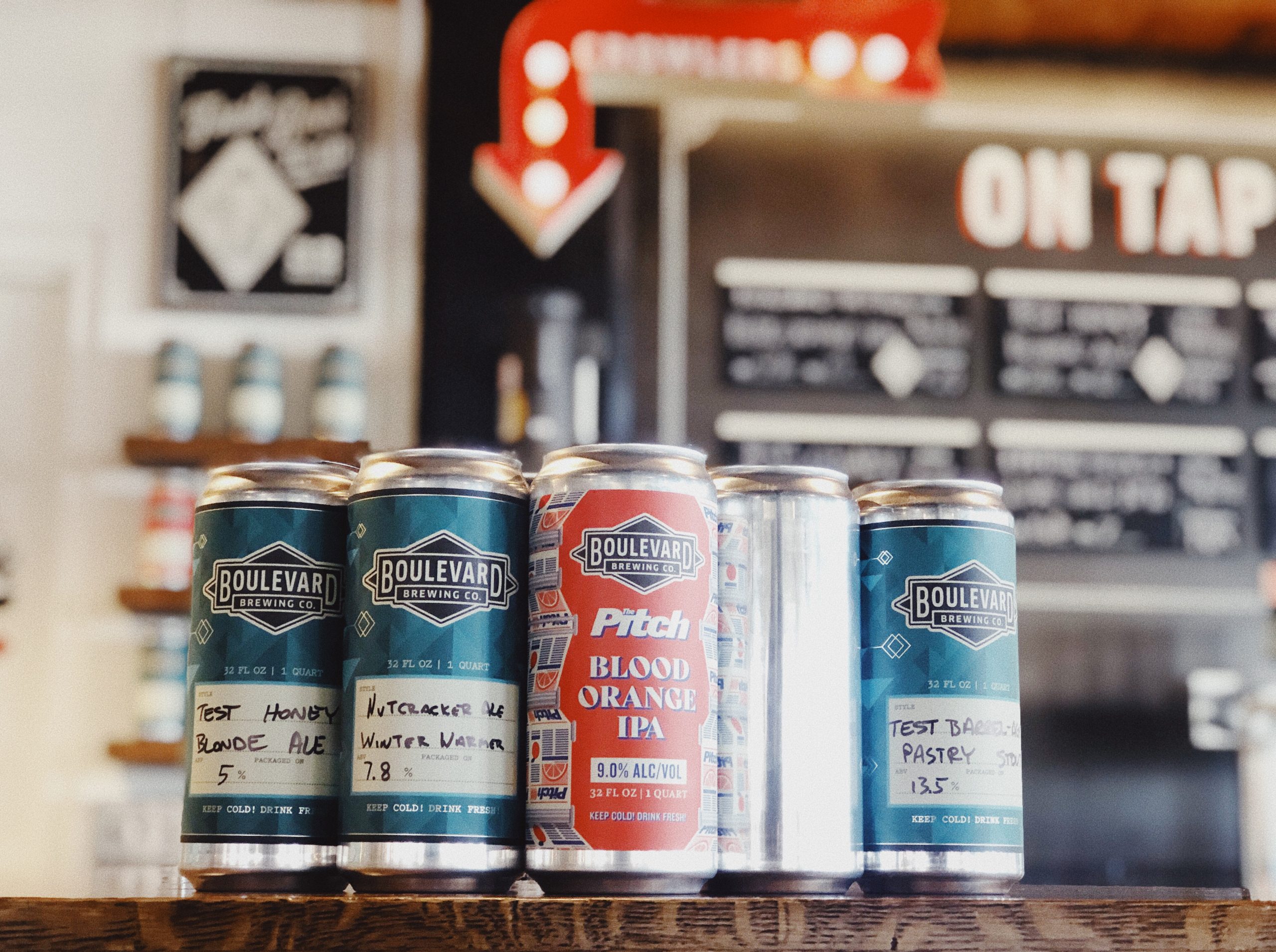 Cans of beer from Boulevard Brewing Co. are lined up on a bar counter. The labels display different types, including a red can of "Pitch Blood Orange IPA." A menu board with tap options is visible in the background.