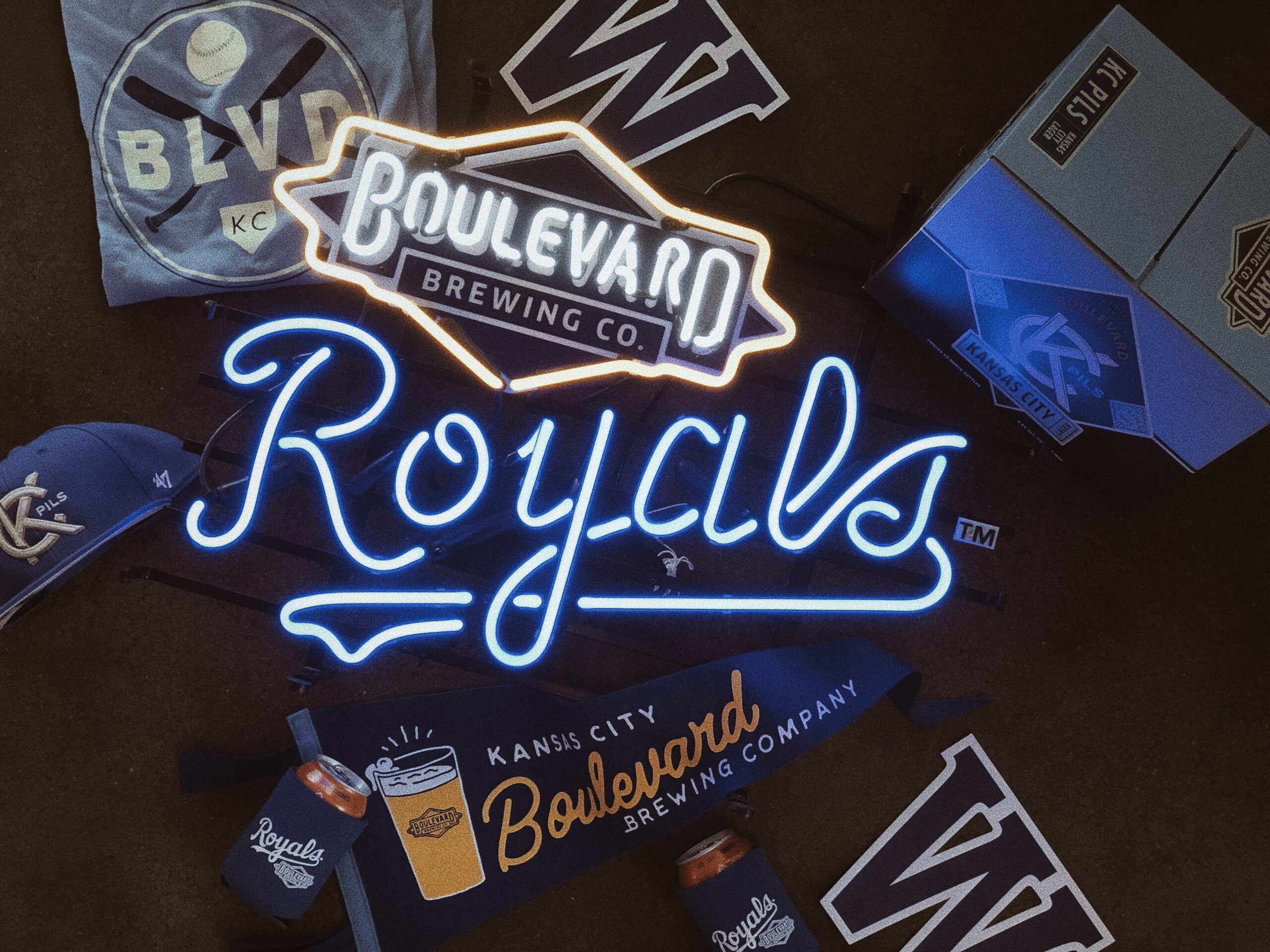 A neon sign reading "Boulevard Brewing Co. Royals" is surrounded by Kansas City Royals merchandise, including T-shirts, koozies with canned drinks, and paper pennants.