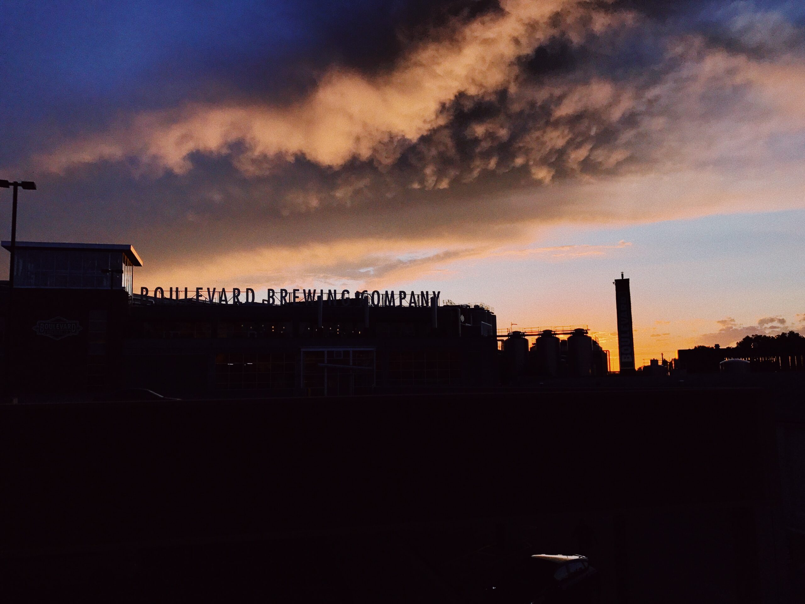 A brewery silhouetted against a dramatic sunset sky with dark clouds. The bold silhouette of the building and its sign contrasts with the colorful hues of orange and purple in the sky.