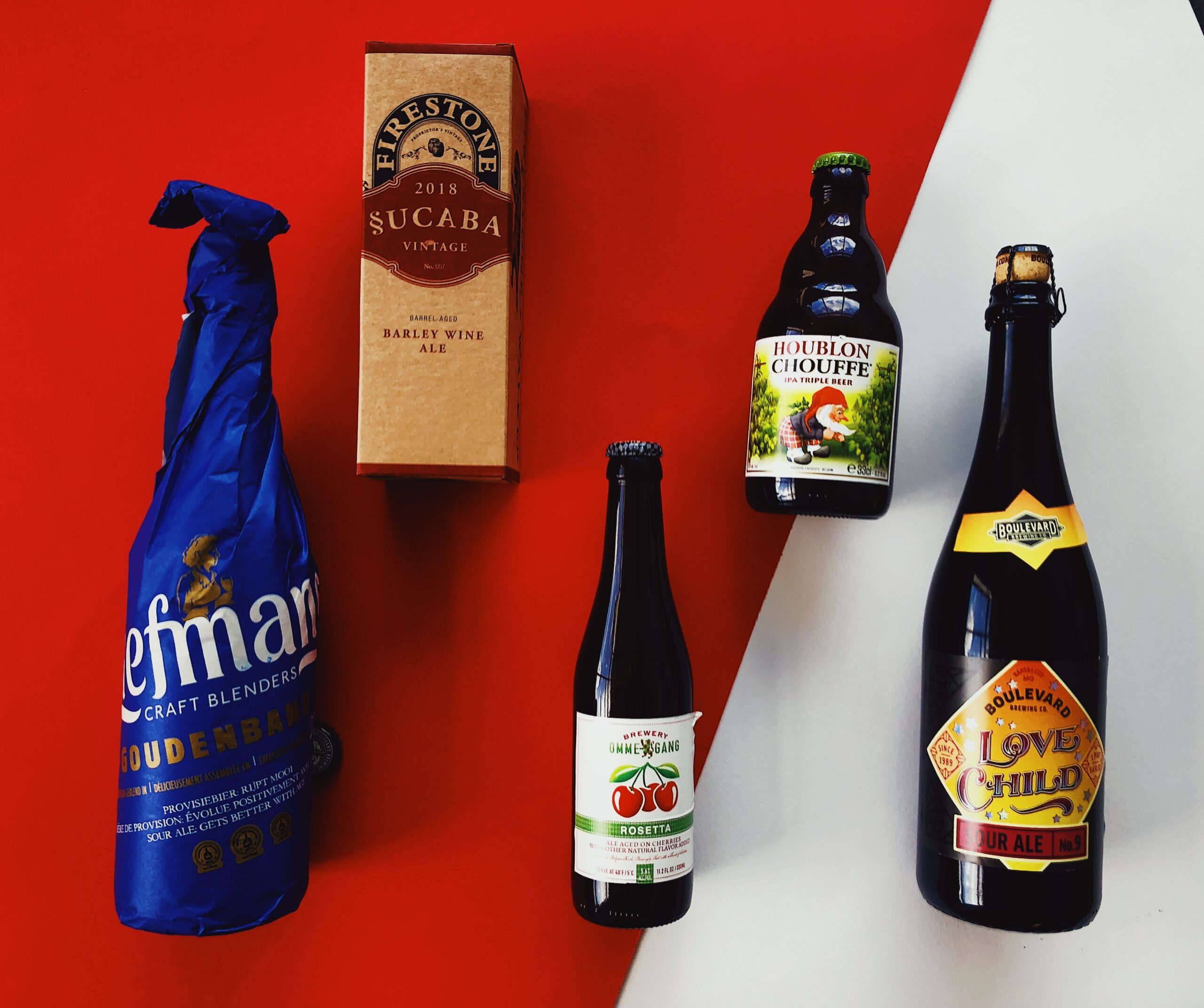 A display of five different beer bottles and boxes arranged on a red and white background. The selection includes various styles and colorful labels, each distinct in packaging design and branding.