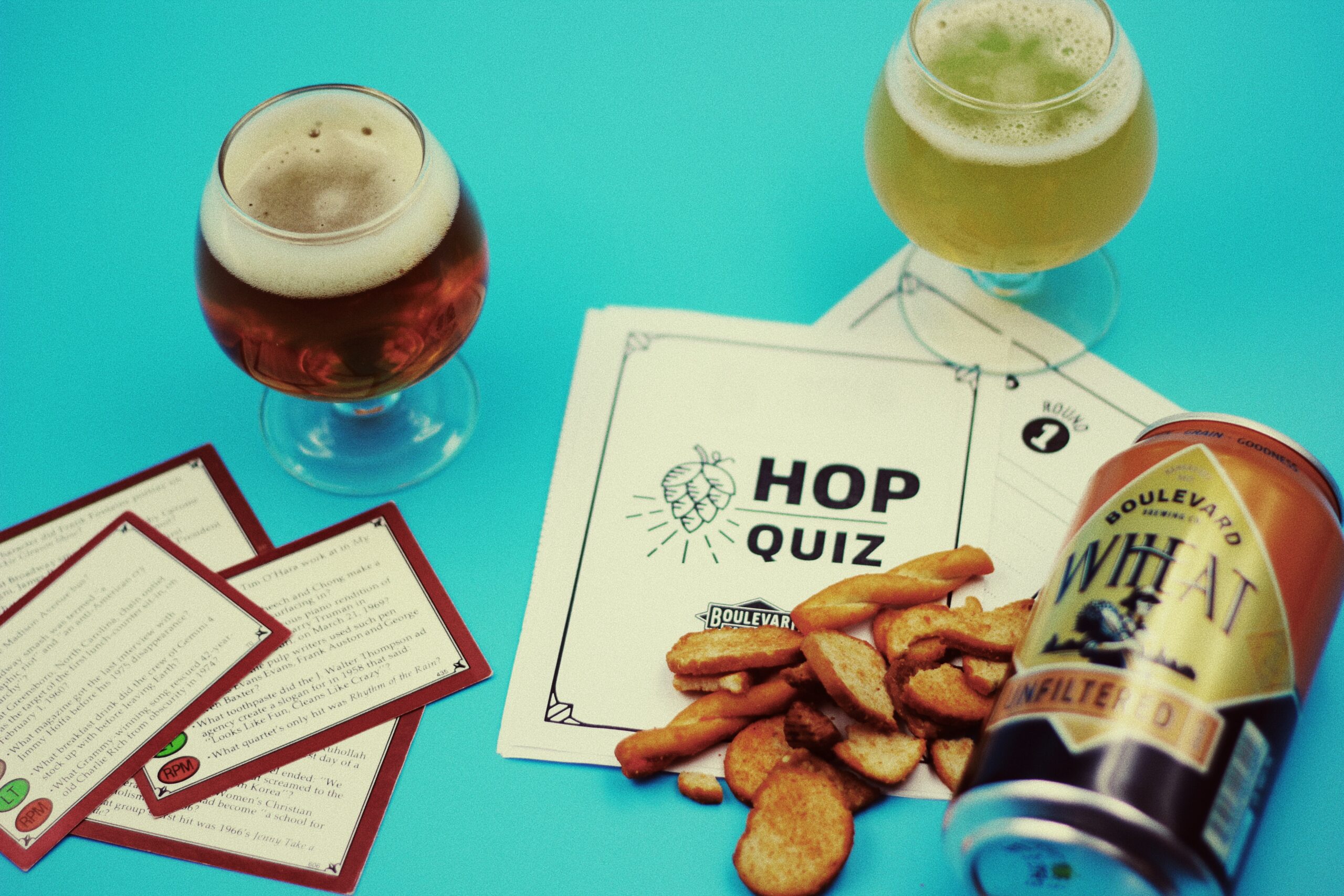 Two glasses of beer, a can of Boulevard Wheat beer, a printed quiz titled "Hop Quiz," and scattered potato chips on a bright blue background.