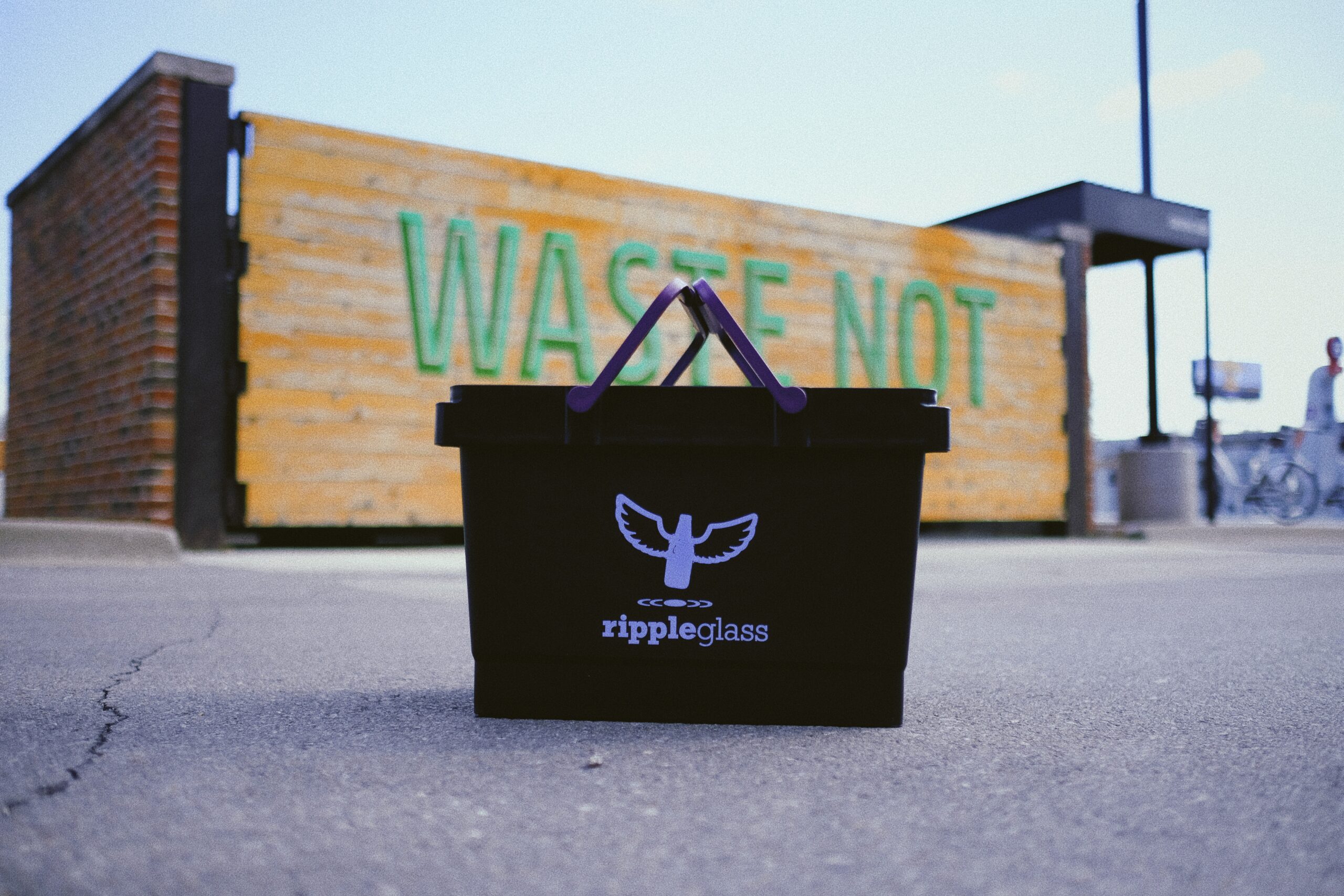 A black recycling bin with "Ripple Glass" and a logo is in focus. In the background, there's a wooden structure with the words "WASTE NOT" in large green letters. The scene is outdoors, with pavement and partial view of a person near a bicycle.