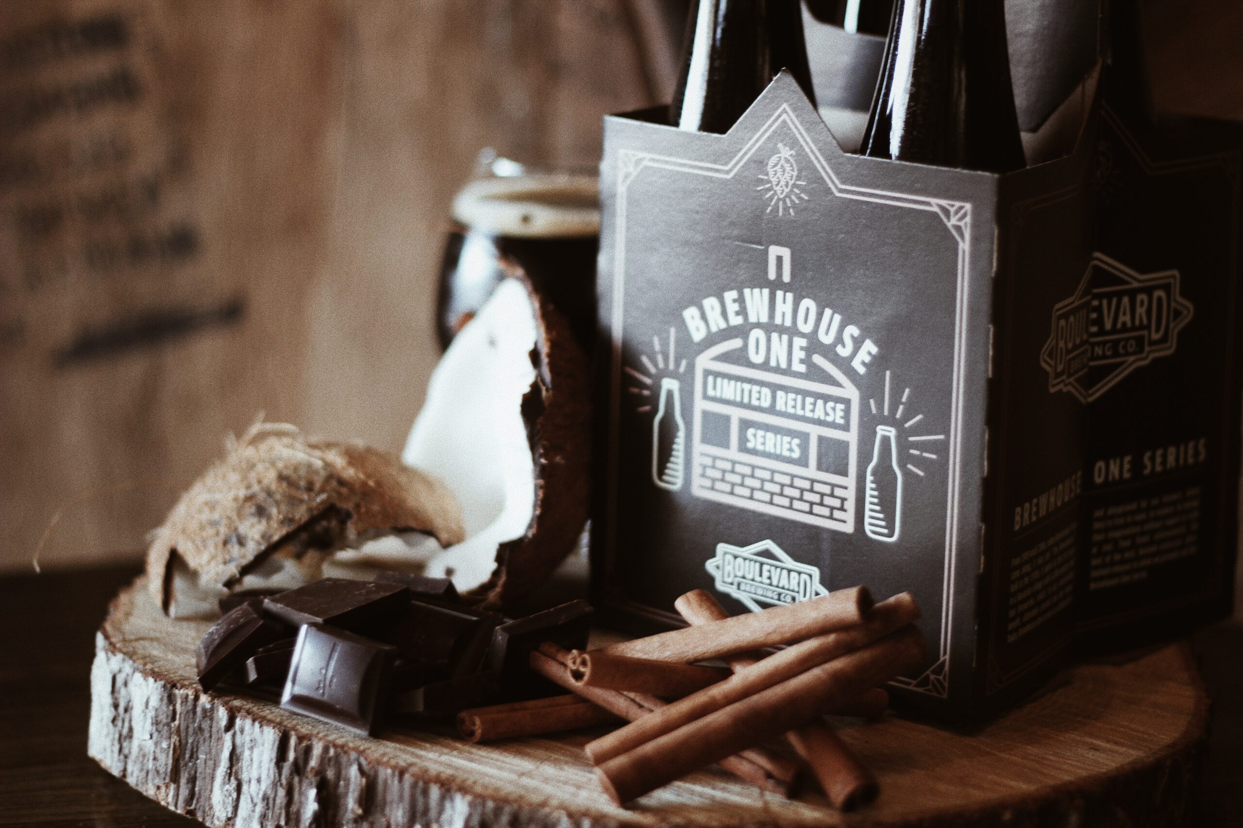 A pack of four Brewhouse One beer bottles sits on a wooden slice with pieces of coconut, chocolate, and cinnamon sticks around it, creating a rustic and inviting display.