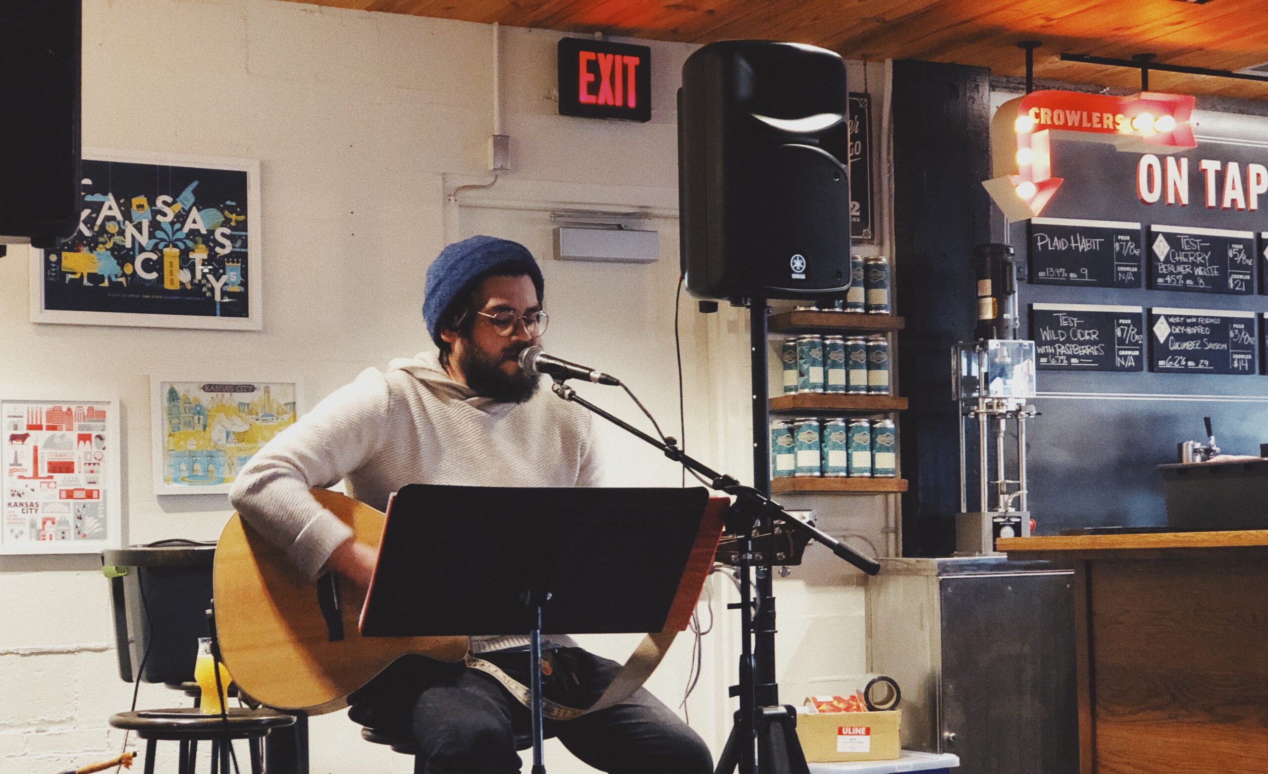 A person wearing a beanie and glasses playing an acoustic guitar and singing into a microphone at a cozy cafe. There are art pieces on the walls and a menu board that reads "On Tap" in the background.