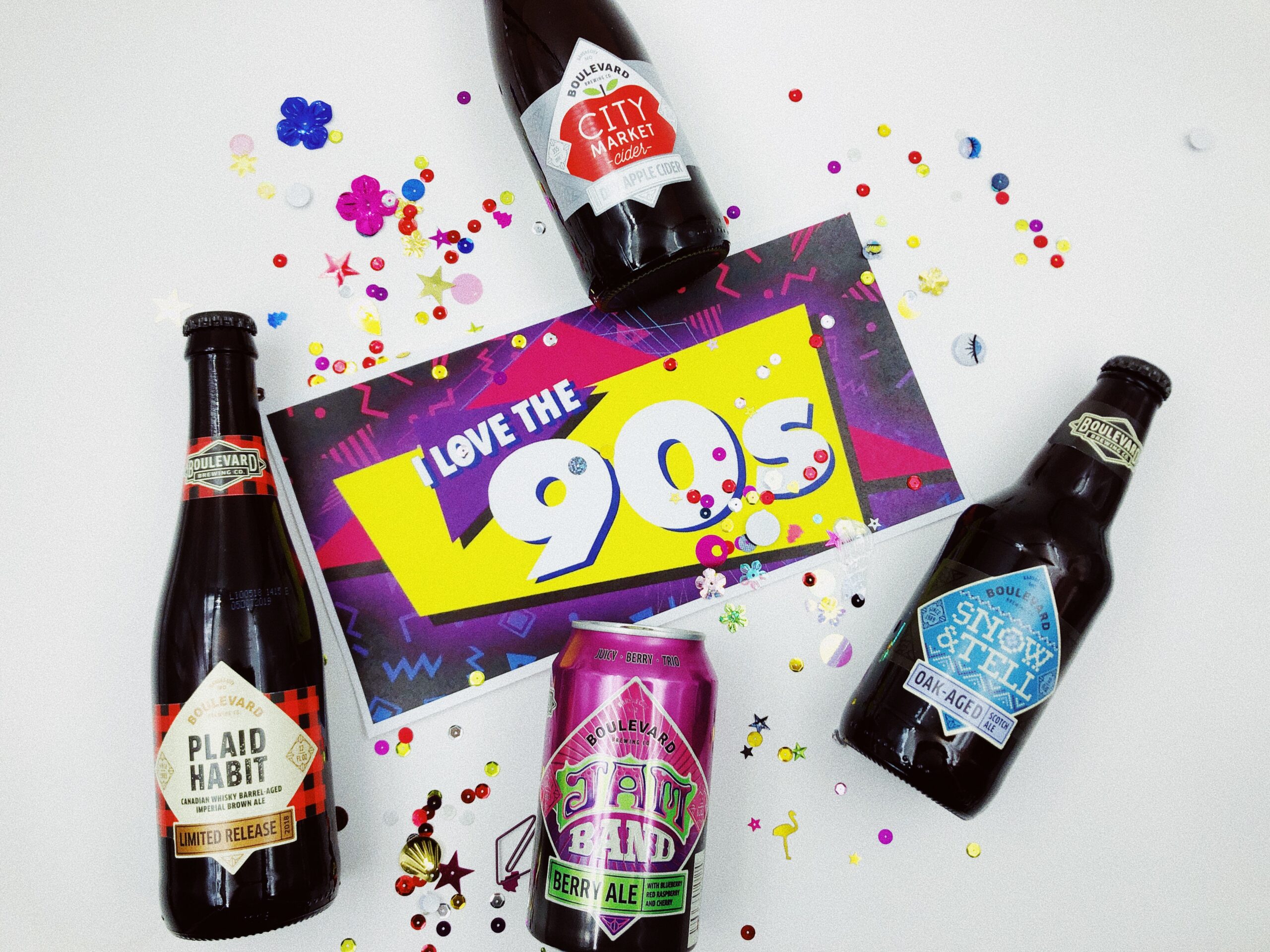 A colorful "I Love the 90s" sign is surrounded by four bottles and cans of craft beer with confetti scattered around them. The beers have retro-style labels.
