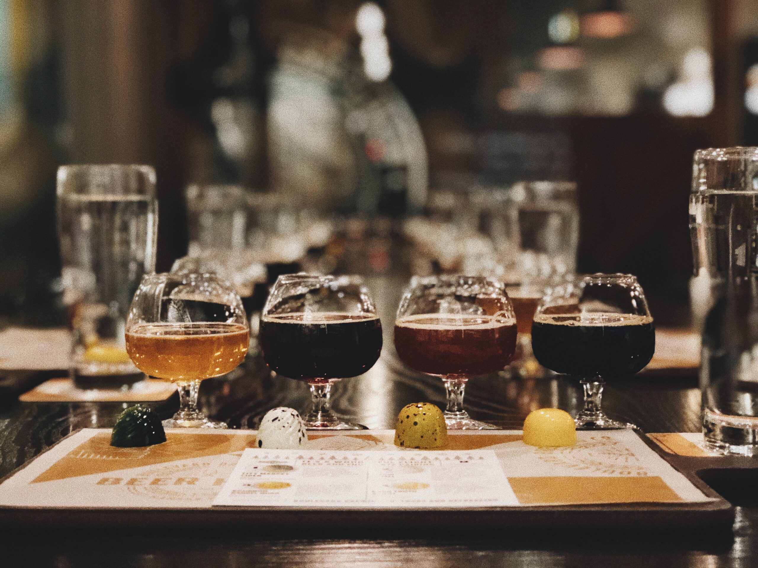 A row of craft beer tasters in small glasses is arranged on a wooden table, each with different colors. The background is blurred, and there's a variety of beverages and water glasses visible.