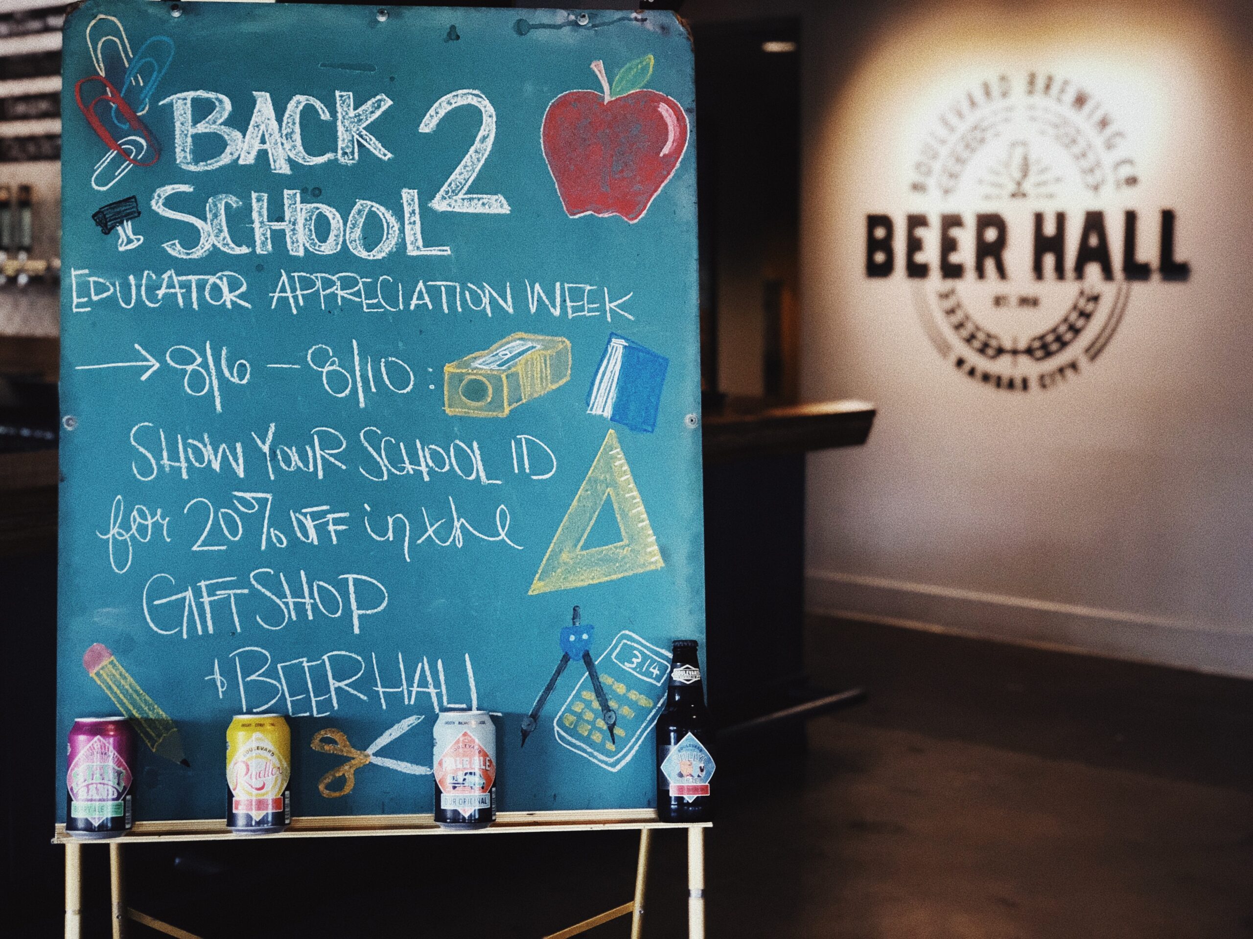 A chalkboard sign reads "Back 2 School Educator Appreciation Week 8/6-8/10: Show your school ID for 20% off in the gift shop + beer hall!" It's decorated with colorful school-themed drawings. Four colorful cans are displayed at the bottom.