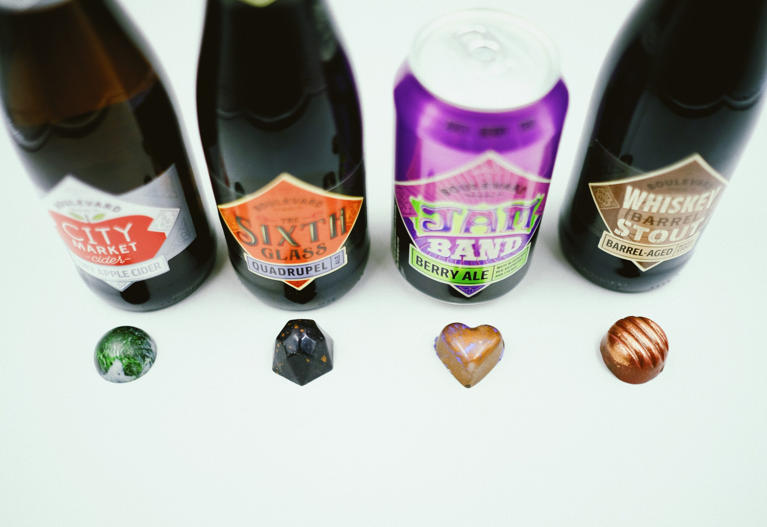 Four beverages are lined up, including bottles and a can, behind four chocolates. The bottles have distinctive labels, and the chocolates are a variety of shapes and colors, placed on a white surface.