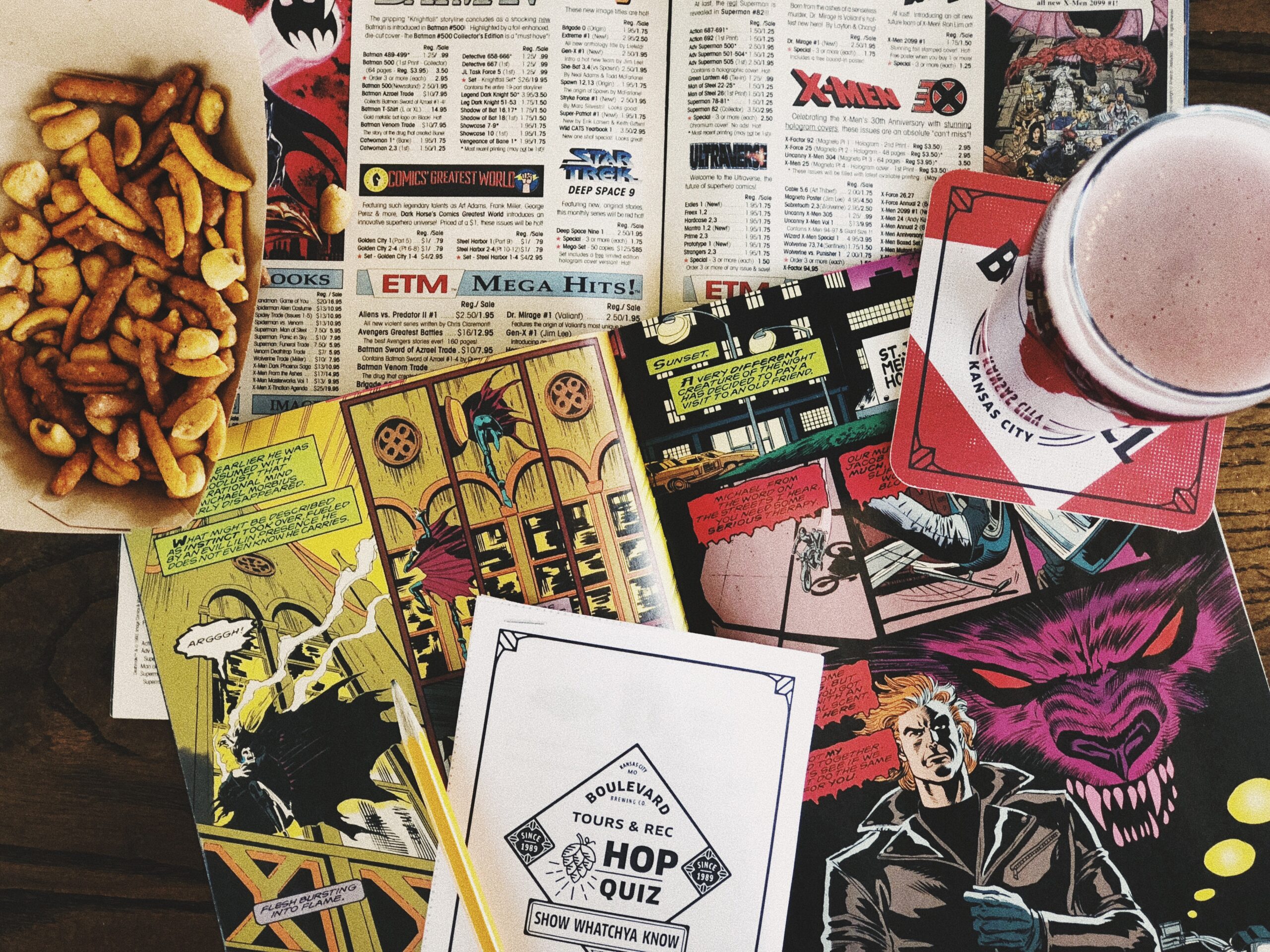 A table with comic books, a newspaper, a bowl of snacks, and a glass of beer on a coaster. The scene captures a cozy, casual setting for reading and enjoying a drink.