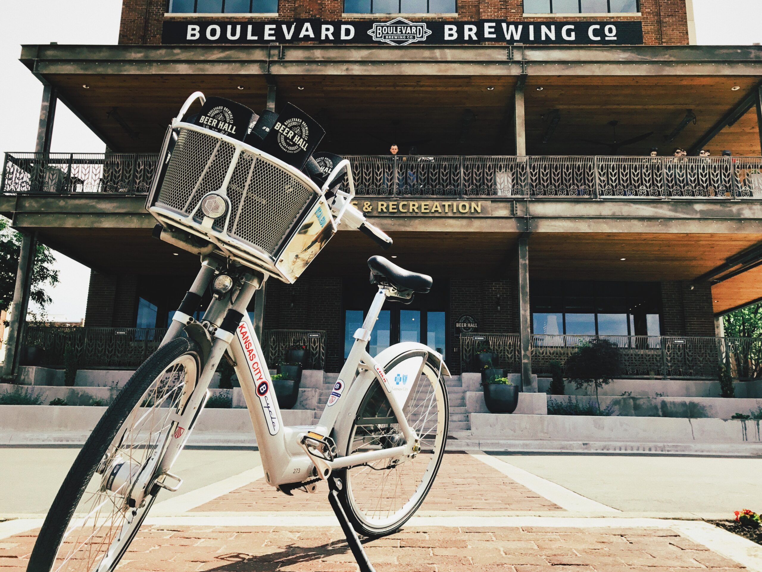 A bicycle with a basket is parked in front of a brewery. The building has signs saying "Boulevard Brewing Co" and "Beer Hall & Recreation." The scene captures a sunny day with clear skies.