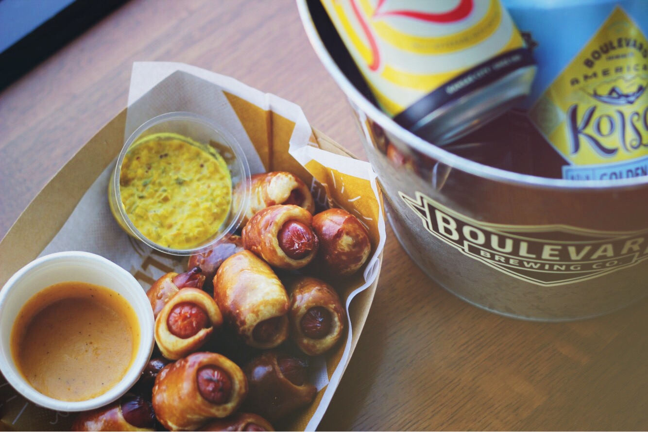 A paper tray with mini pretzel-wrapped hot dogs is accompanied by a cup of mustard dip and another dipping sauce. Nearby, a metal bucket holds canned beverages, branded with Boulevard Brewing Co.