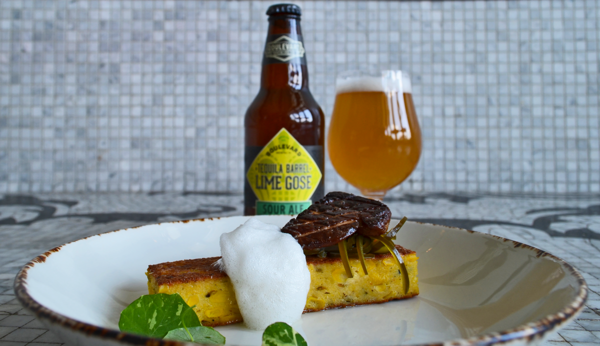 A dish featuring a block of cornbread topped with garnishes and foam sits on a plate. Behind it, a bottle of tequila barrel lime gose and a filled glass are displayed on a patterned surface.