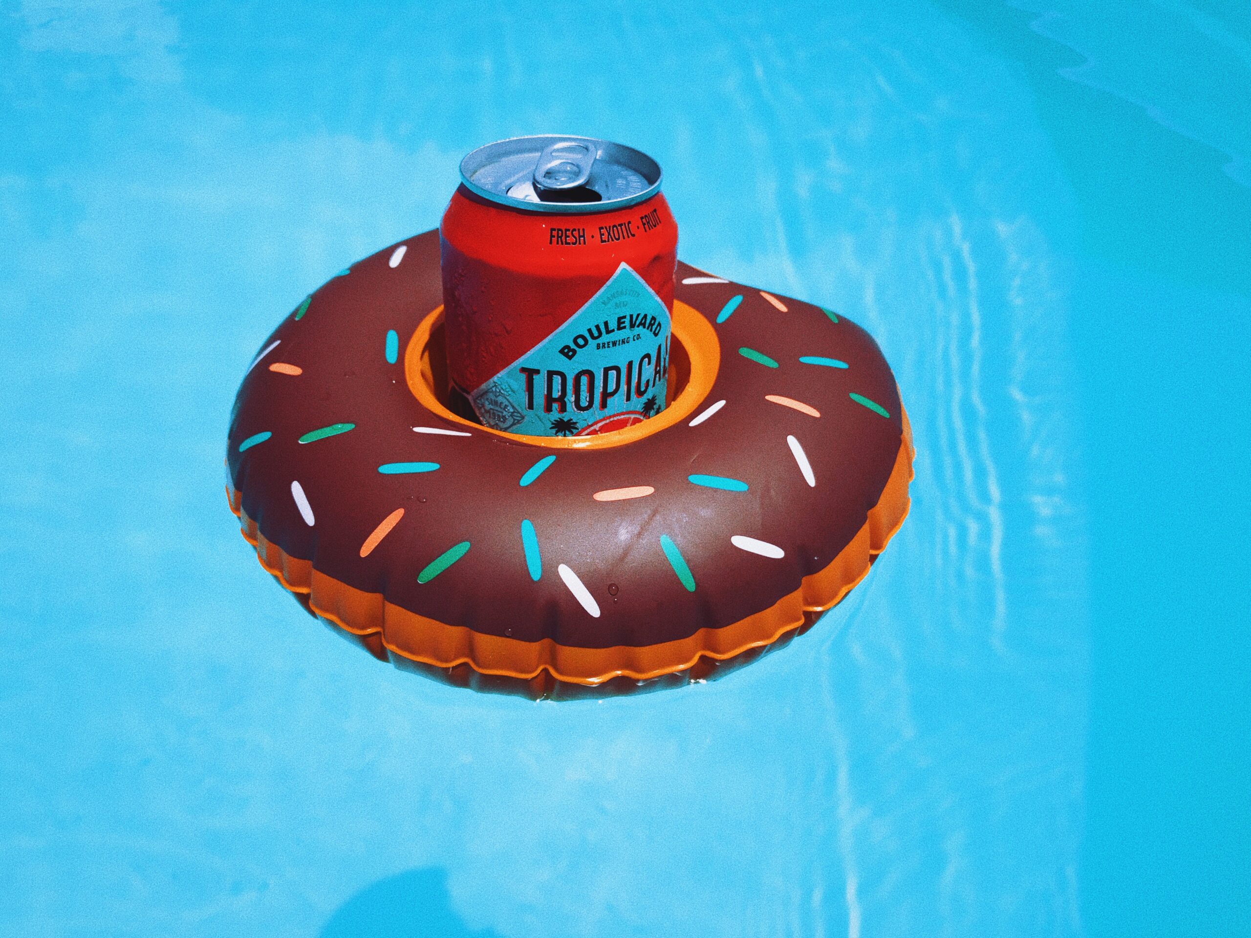 A can of Boulevard Tropic beer placed in a donut-shaped pool float is floating on clear blue water. The float is designed to look like a chocolate donut with colorful sprinkles.