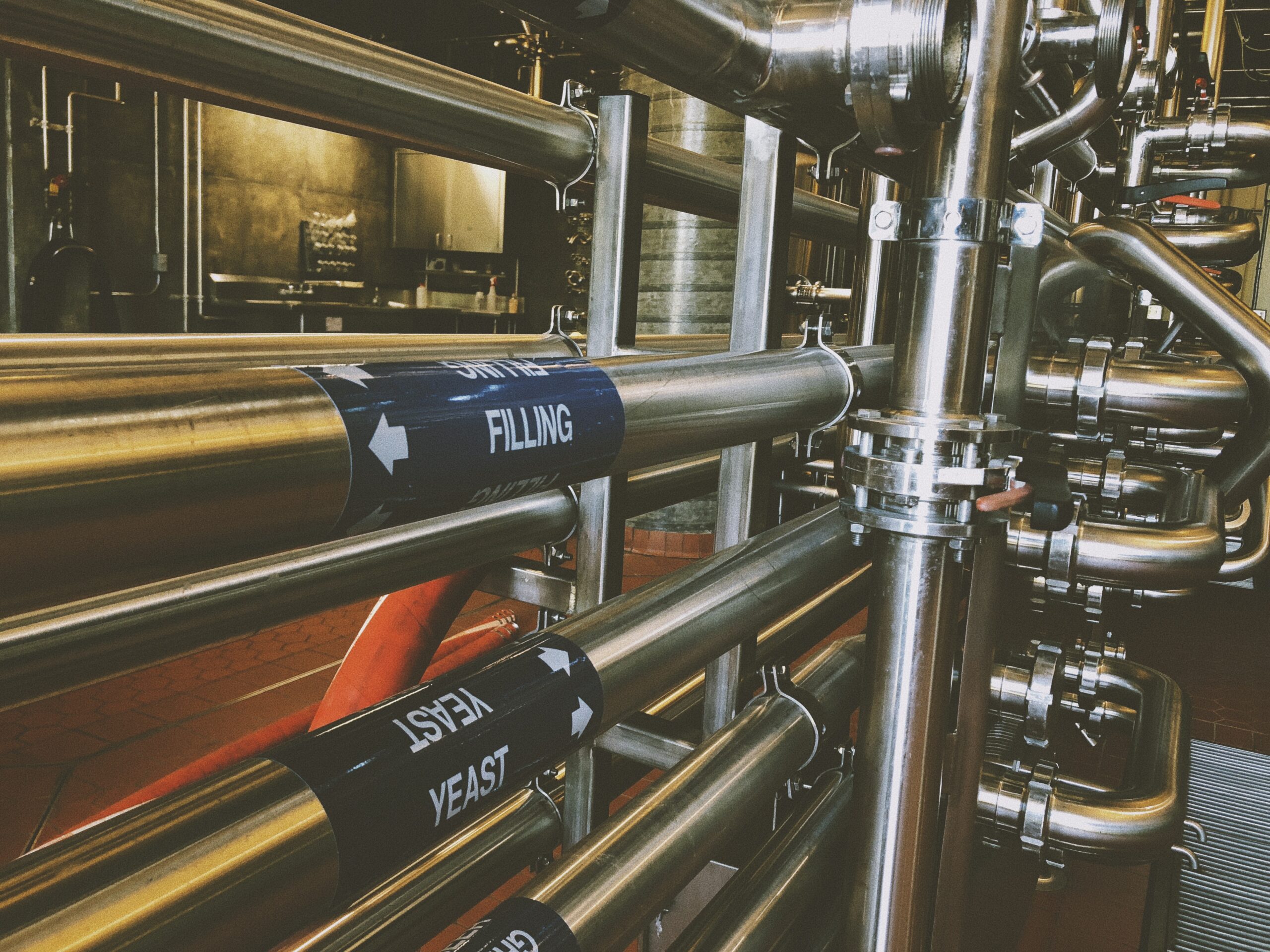 A complex arrangement of stainless steel pipes in a brewery, with labels indicating directions for "Filling" and "Yeast". The background features an industrial setting with additional equipment and dim lighting.